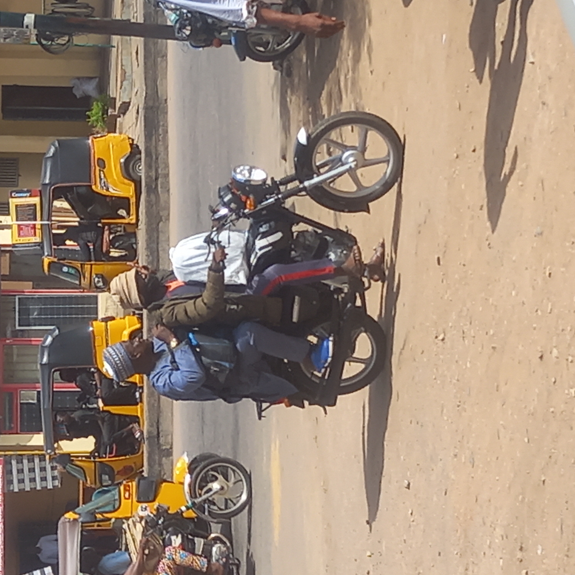 The image shows three men riding a motorcycle on a street in Dadi Riba, Nigeria. The motorcycle is black with silver wheels. The first man, wearing a blue shirt, sits in front and appears to be driving. The second man is wearing a tan head covering and a green jacket. The third man is in the middle. All are dressed in casual clothing. The motorcycle is traveling on a paved street, and several yellow auto rickshaws are parked nearby. The background shows buildings and a pole with signage. The lighting suggests it is daytime.