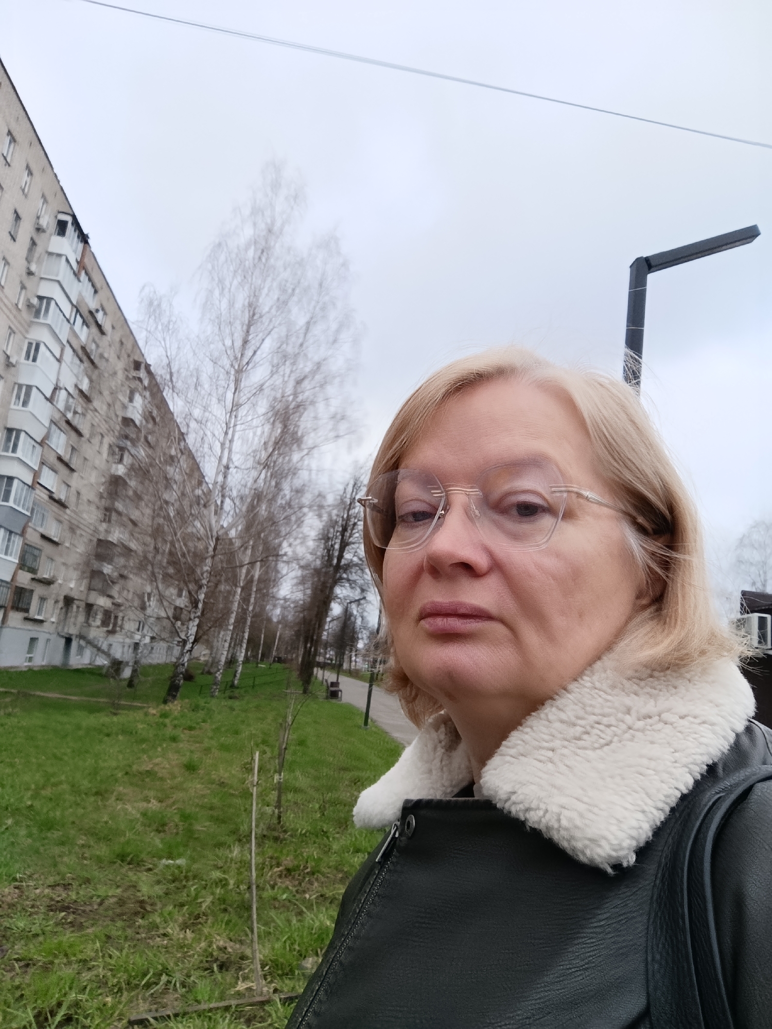 The image is a medium shot of a woman outdoors. She has short, blonde hair and is wearing glasses, a dark-colored leather jacket with a white fur collar, and a black shoulder bag. She is looking directly at the camera with a neutral expression.
Behind the woman is an urban landscape. To the left is a multi-story apartment building with a repetitive pattern of windows and balconies. In front of the building are bare trees lining a grassy area that appears to be a park or green space. There is a sidewalk or pathway visible in the background, as well as a dark-colored lamppost on the right.
Based on the visual information available, the location is likely Chaikovsky, Russia, as per the prompt's request. The architecture of the apartment building is consistent with Soviet-era construction common in many Russian cities.