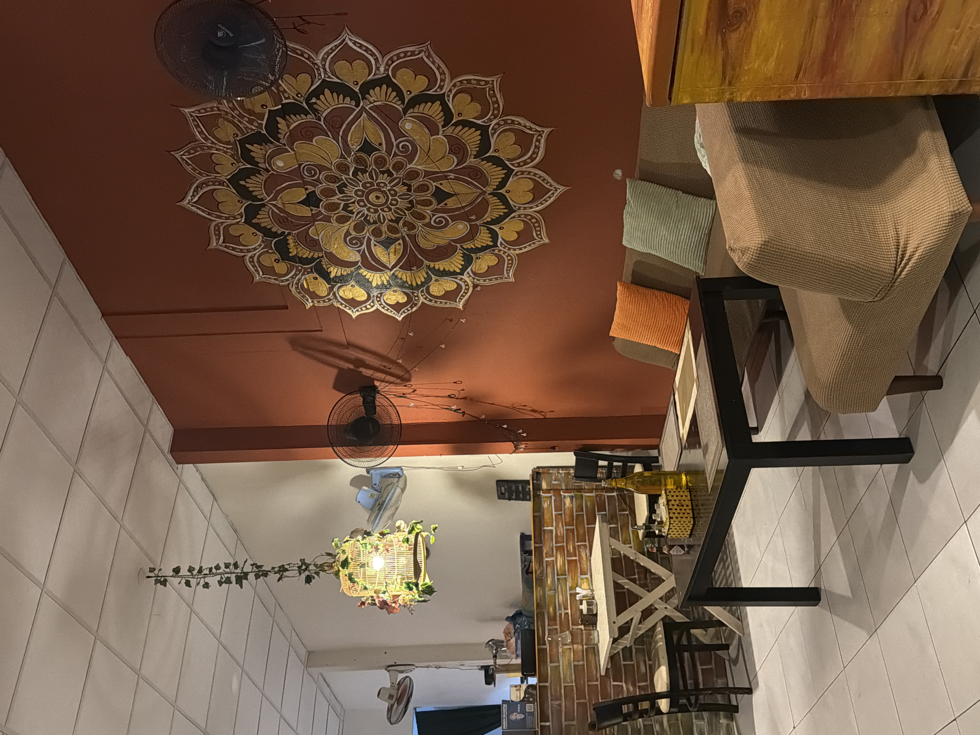 The image shows an indoor cafe setting. In the foreground, a dark rectangular table is set with placemats, a bottle of yellow liquid, and what appears to be a small basket with items inside. To the left of the table are two chairs with cushioned seats, and a small white table with a checkered tablecloth. Beyond this, there's a brick wall. In the background, a brown sofa with two colorful cushions (orange and light green) is visible. To the right, a wooden cabinet with a colorful, possibly painted, wood-grain pattern is present. The walls are painted a warm terracotta color, and a large, circular, mandala-like painting is prominently displayed on the right wall. There are also fans mounted on the walls and a decorative birdcage hanging from the ceiling, adorned with artificial flowers and greenery. The floor is tiled. The overall ambiance suggests a relaxed and possibly ethnic-themed cafe. The provided location is Bac Nha Trang Ward, Vietnam, but this cannot be visually confirmed from the image itself.