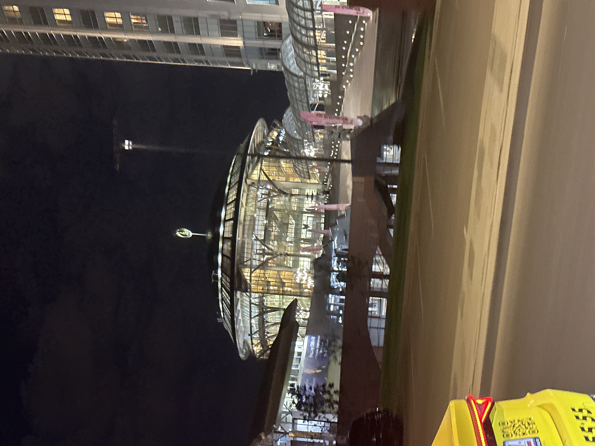 The image shows the Crystal Island complex in Moscow, Russia, at night. The complex is distinguished by its multi-tiered glass and steel structure, illuminated with interior lighting. A tall building is visible in the background. A sculpture stands in front of the building. A yellow advertisement structure is in the foreground. There are no people visible in the image. The sky is dark, indicating nighttime.