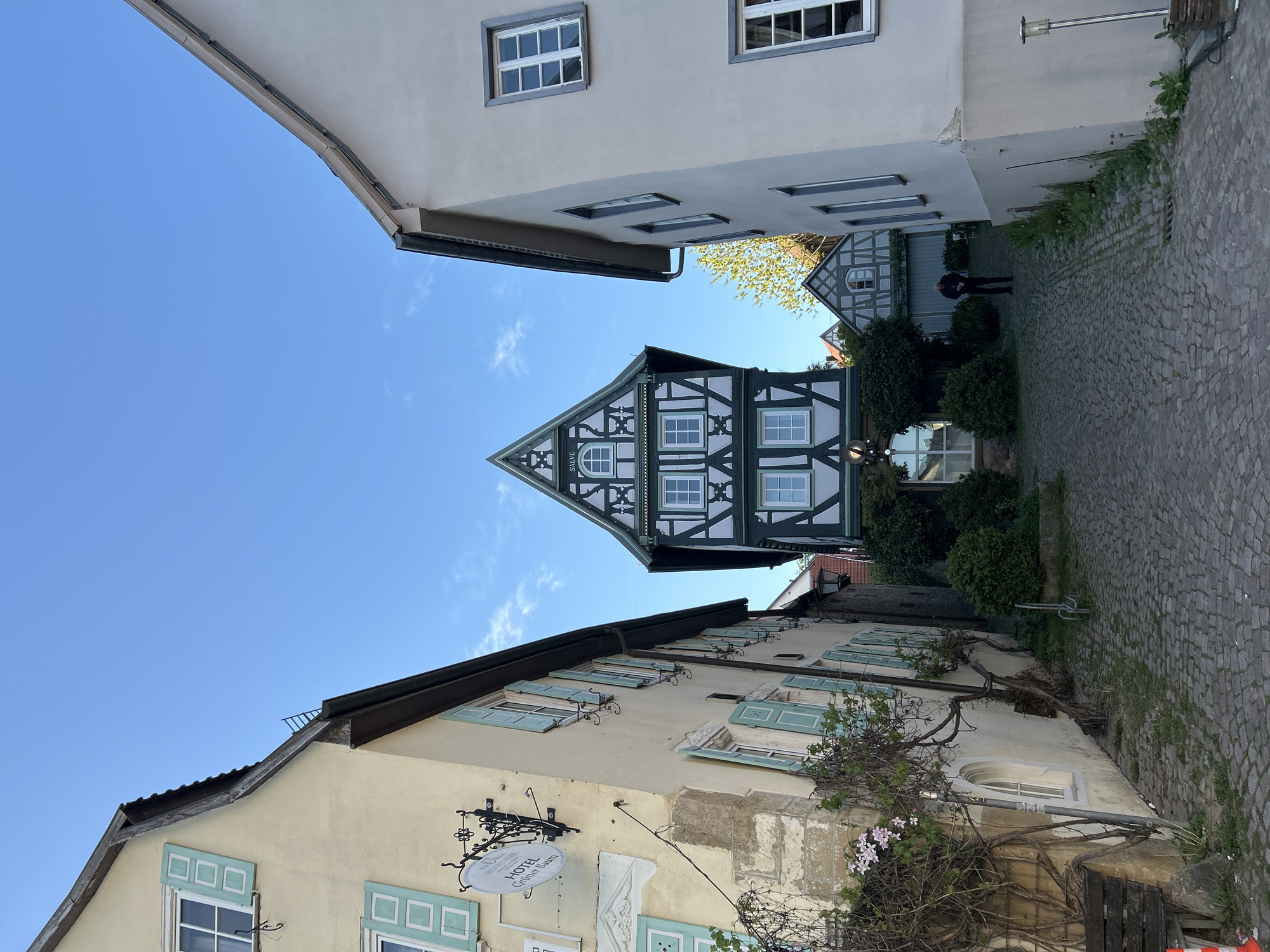 The image captures a narrow cobblestone street between buildings in an unknown city. The sky is blue. On the left, a building with light yellow walls and light blue window shutters is visible. A sign for the "Hotel Gruner Baum" is attached to the building's exterior. On the right, a lighter-colored building also runs along the street. Straight ahead, framed by the two buildings, is a half-timbered building with a triangular peak. A person can be seen in the distance on the cobblestone path. Shrubs and plants grow along the edge of the pathway.