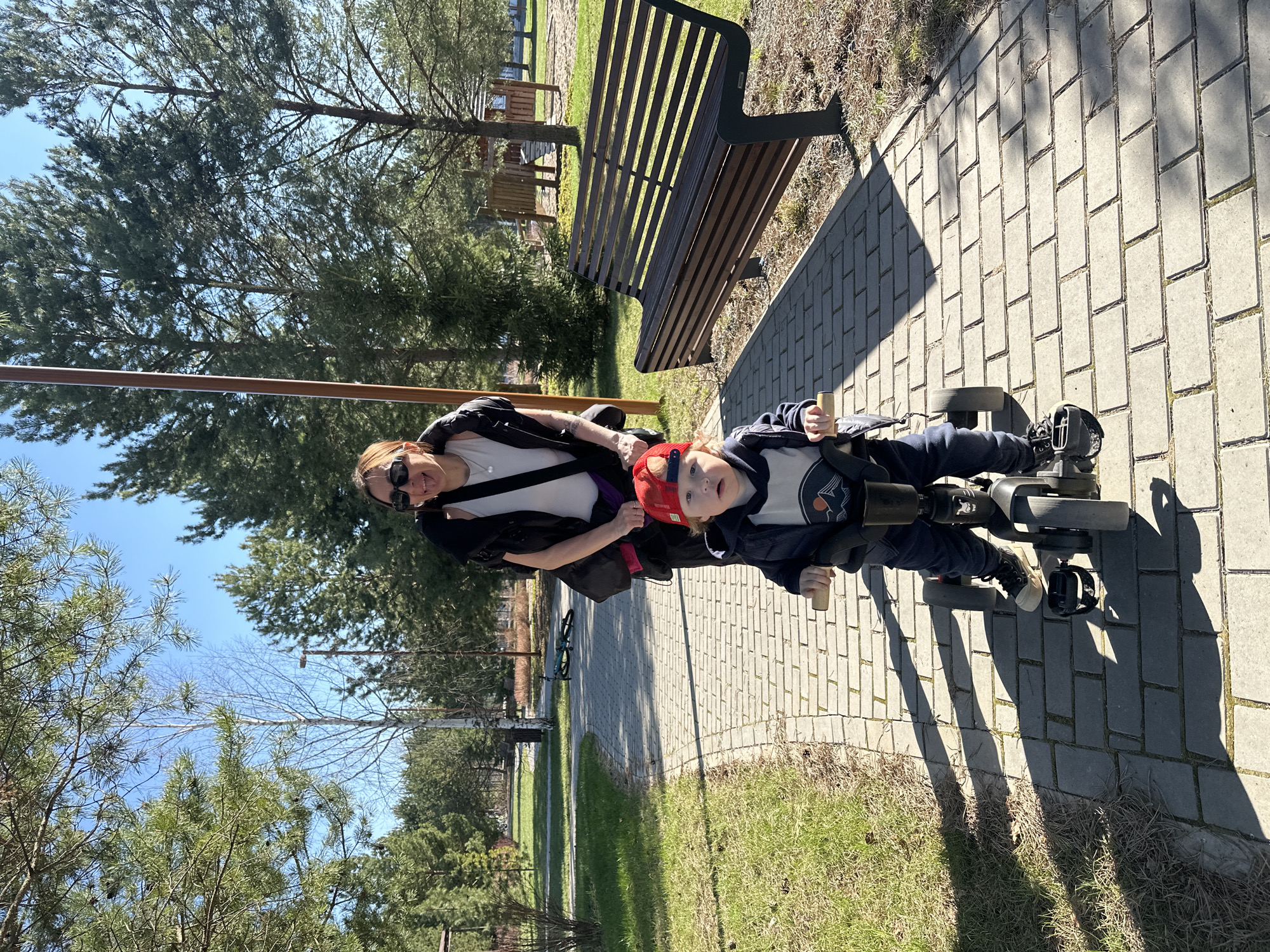 The image depicts a woman with sunglasses standing beside a child on a tricycle or adaptive bike. The child wears a red cap, and both are on a paved pathway. To the right of the pathway, there's grass. Behind them is a wooden park bench and trees. The sky is visible in the background. This scene is located in a park in Русский Вожой, Russia.