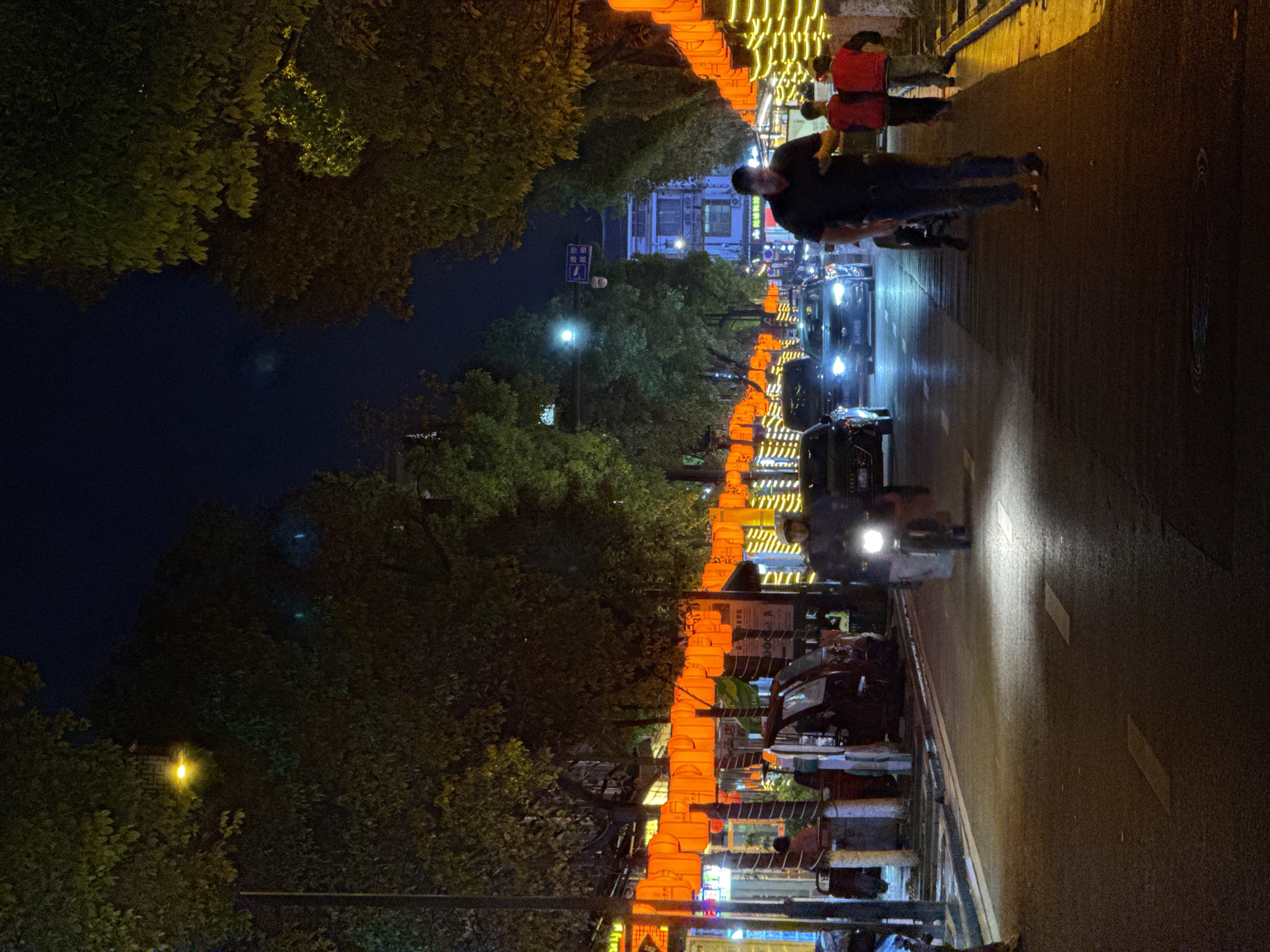 The image depicts a street scene at night in Wulingyuan, China. The street is lined with orange lanterns and decorative lights. People are walking along the sidewalk. Cars and scooters are driving on the street. Trees are on the side of the road.