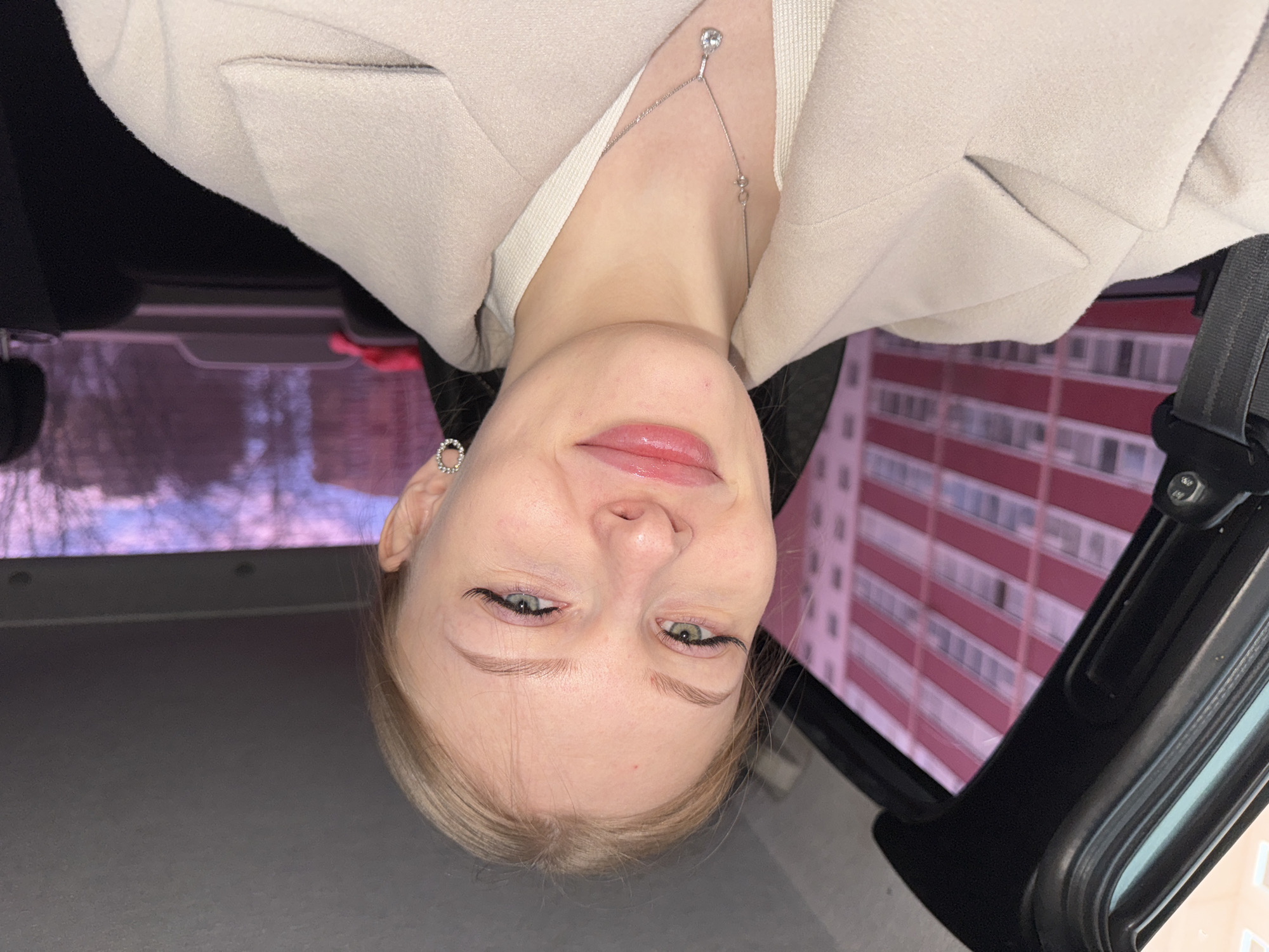 A young woman is in a car, wearing a light-colored blazer and a delicate silver necklace. Outside the car window is a tall apartment building with horizontal red and white stripes. The image was taken in Perm, Russia.