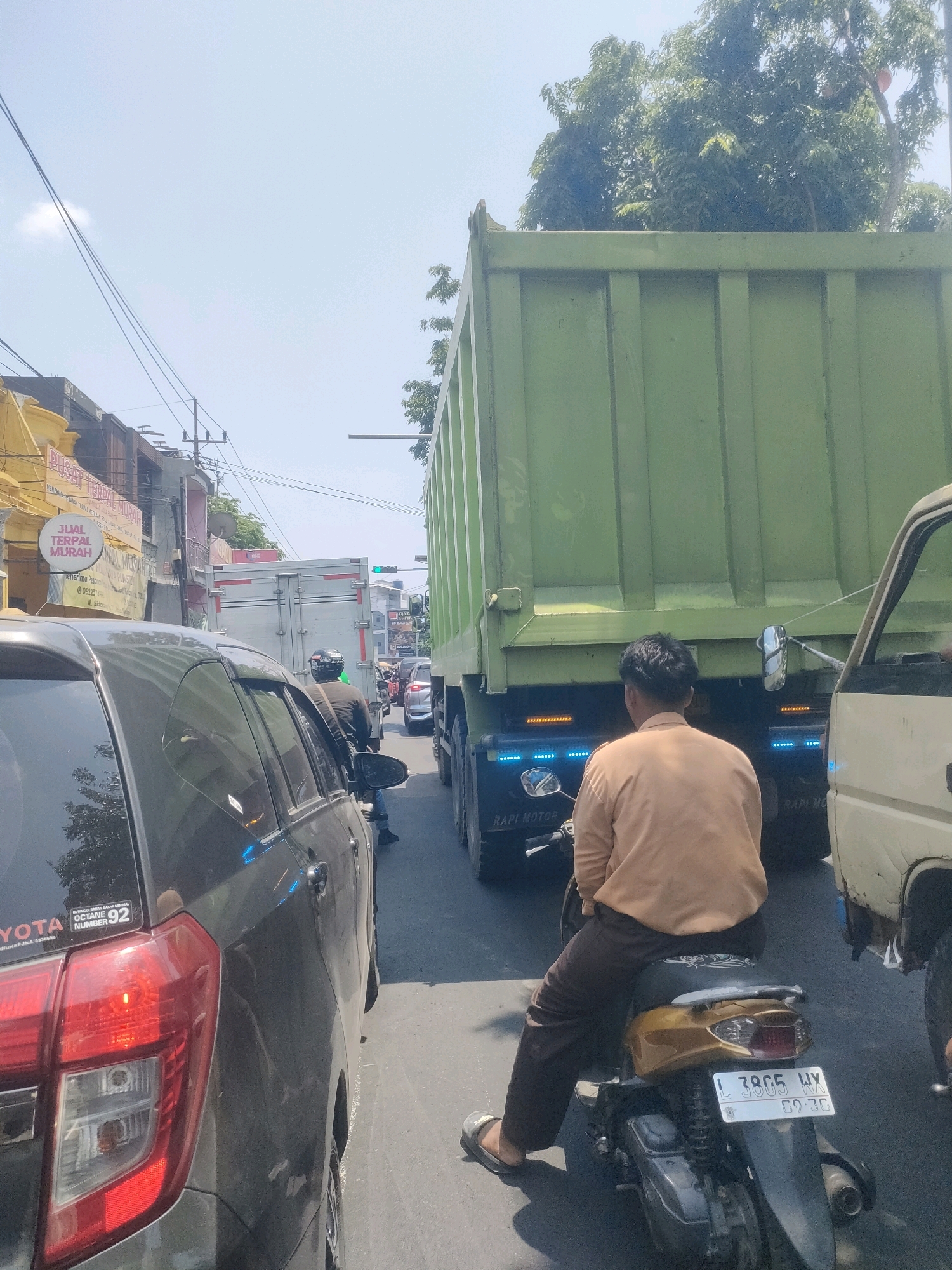 The image depicts a traffic jam on a sunny day in Surabaya, Indonesia. In the foreground, a dark-colored car is partially visible on the left, with its taillight illuminated. To its right, a person wearing a light brown shirt and dark pants is riding a gold and black scooter, facing away from the camera. The scooter has a license plate with the characters "L 3805 WX". Behind the scooter, a large green dump truck dominates the center of the image, and to its right, a section of a cream-colored truck is visible. In the background, a white truck is stopped in traffic, and further down the road, a traffic light shows a green signal. Buildings line the left side of the street, with signs indicating shops and businesses, including one that reads "PUSAT TERPAL MURAH" (Cheap Tarpaulin Center). Electrical wires crisscross overhead. The scene is characteristic of urban traffic congestion in Indonesia.
