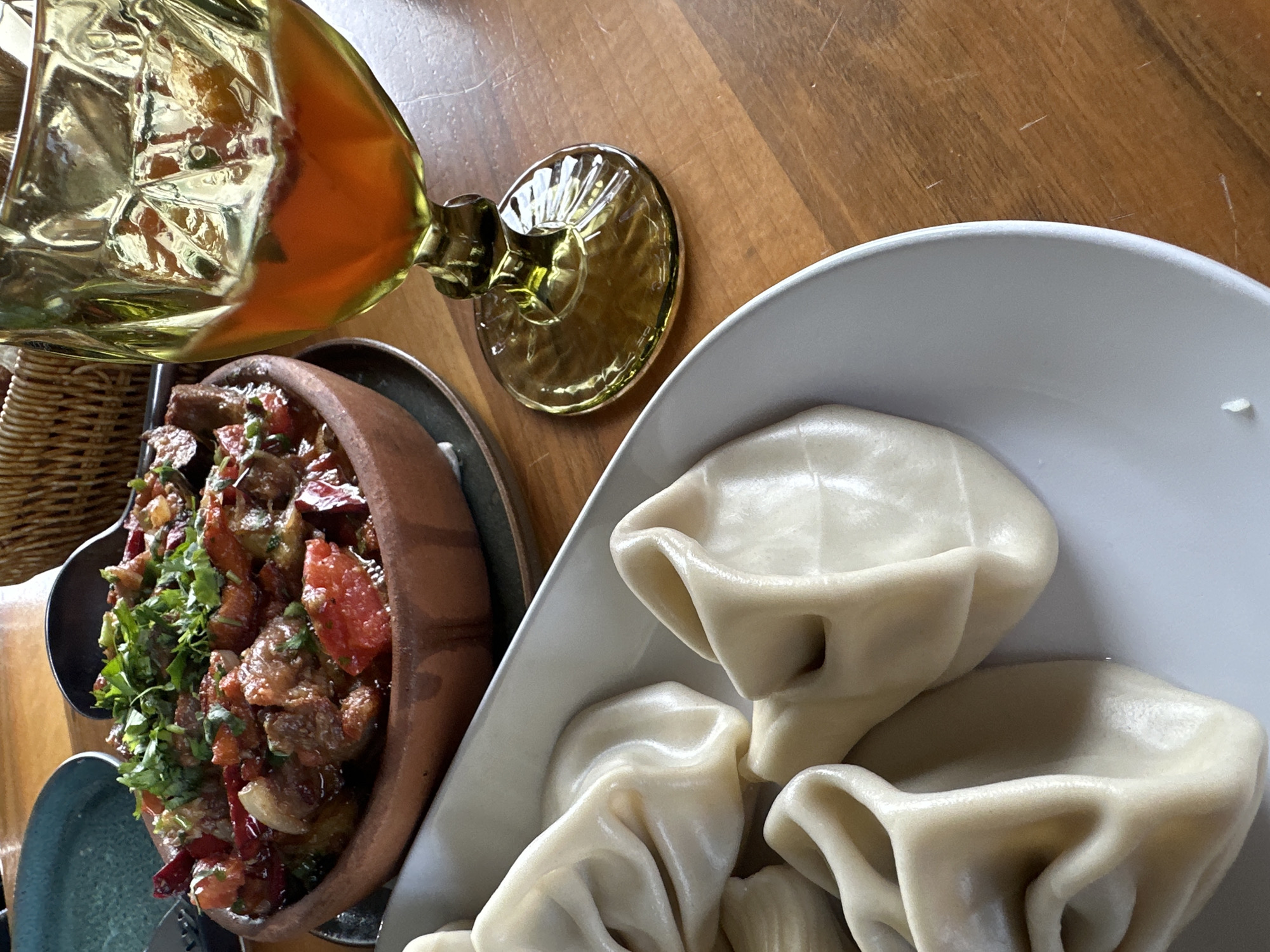 A high-angle shot shows two dishes on a wooden table, alongside a large golden glass.
The dish on the left is a dark-colored clay pot containing a stew with pieces of meat, tomatoes, peppers, and chopped herbs. The pot sits on a metal saucer, and a blue dish and cutlery are visible to the left.
To the right, a white plate holds several large dumplings.
The location is Marneuli, Georgia, as indicated by the food being served in a restaurant. No people are visible in the frame. The setting appears to be indoors, given the wooden table.