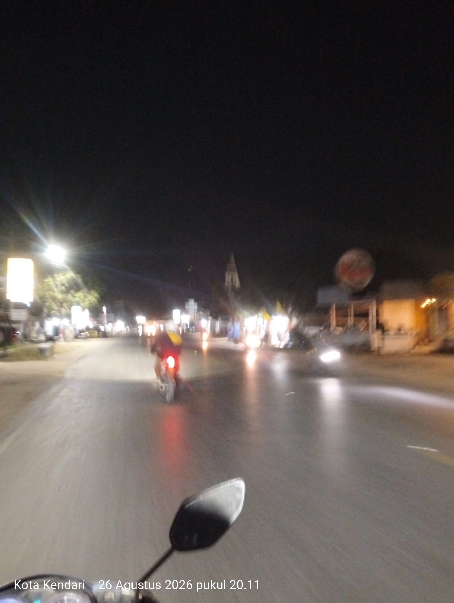 The image is a night-time photograph taken from a first-person perspective, likely from a motorcycle, showing a street scene in Kendari, Indonesia on August 26, 2026, at 8:11 PM. The road is wet, reflecting the lights from street lamps and passing vehicles. A motorcycle is visible in the middle distance, with its taillight illuminated, moving away from the camera. Other vehicles and indistinct figures are present along the sides of the road, suggesting a moderately busy street. Buildings and illuminated signs line the sides of the street, with trees visible in the background. The overall impression is of a typical urban street environment at night, with the characteristic blurred motion associated with a moving camera.