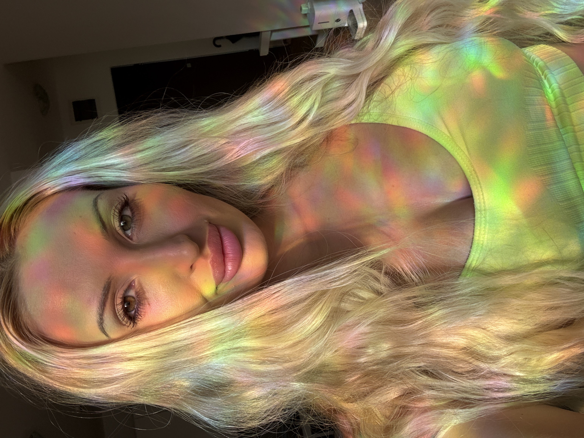 A blonde woman with long, wavy hair is photographed from the chest up in a close-up shot. She is wearing a light green, ribbed tank top. The lighting in the image is dynamic, with multicolored, prismatic light patterns casting across her face, hair, and upper body, creating a vibrant and ethereal effect. The background is dimly lit and out of focus, with hints of what appear to be indoor furniture and a doorway. The woman's expression is neutral, with her gaze directed slightly off-camera.