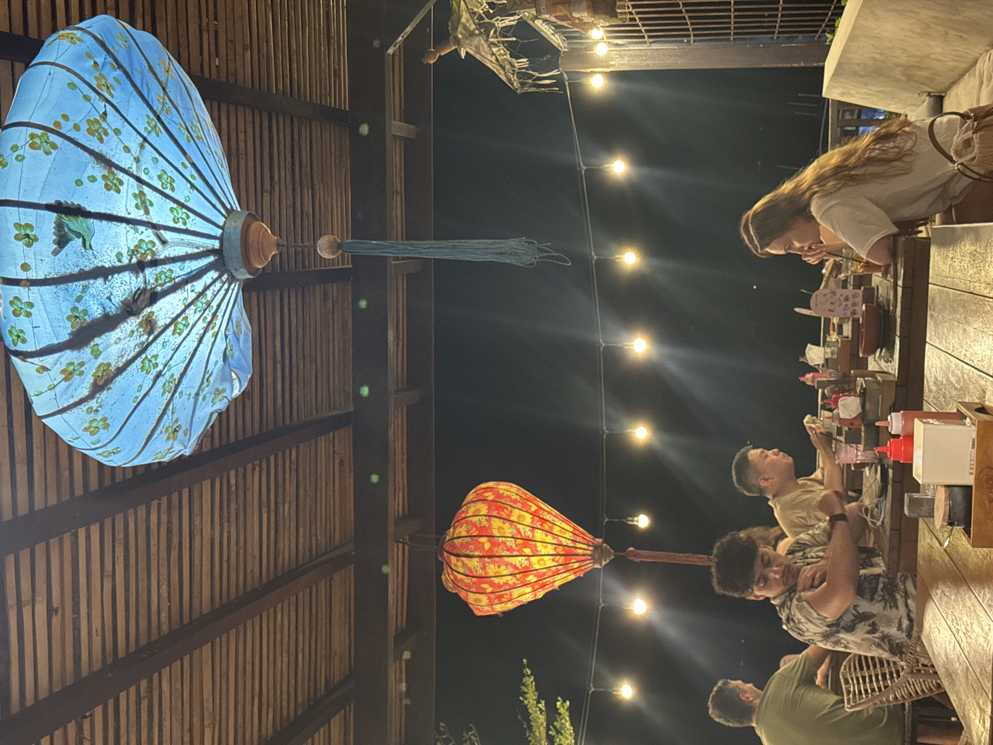 The image depicts a nighttime scene at what appears to be an outdoor or semi-open-air restaurant or dining area in Nusa Penida, Indonesia. The setting is dimly lit by artificial light sources, including a string of exposed light bulbs hanging across the frame and two decorative lanterns.
Several people are seated at a long wooden table, engaged in dining or socializing. At the far end of the table, a woman with long hair is visible, leaning over the table. Further down, three men are seated, one in a patterned shirt looking towards the camera, and two others also focused on their meals or companions. The tables are set with various dishes, condiments, and drinks.
Prominently featured are two large, ornate lanterns hanging from a wooden slatted ceiling structure. One lantern, positioned on the left, is light blue with intricate floral patterns, resembling an open decorative umbrella or fan. The other, closer to the center-right, is red and yellow with patterns, shaped like a elongated traditional lantern. Both lanterns are illuminated from within, casting a soft glow.
The ceiling itself is made of wooden slats, giving the area a rustic and natural aesthetic. The dark sky in the background confirms it is nighttime. The overall atmosphere appears casual and relaxed, indicative of a pleasant evening meal or gathering. There is no discernible text visible in the image.