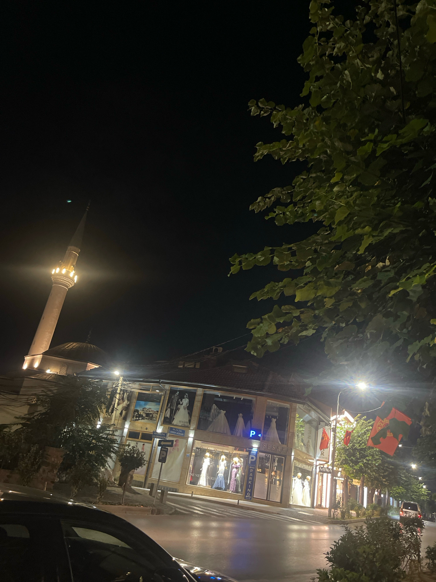 A nighttime street view in Prizren, Kosovo, features a prominent minaret of a mosque illuminated by lights, with its dome visible below. Adjacent to the mosque, a building with large display windows showcases wedding dresses on mannequins. Streetlights illuminate the scene, reflecting on the wet asphalt of the road. Red flags, likely Albanian flags, hang from poles along the street. A black car is parked in the foreground, and a few trees with green leaves are visible. The sky is dark, indicating it is nighttime.