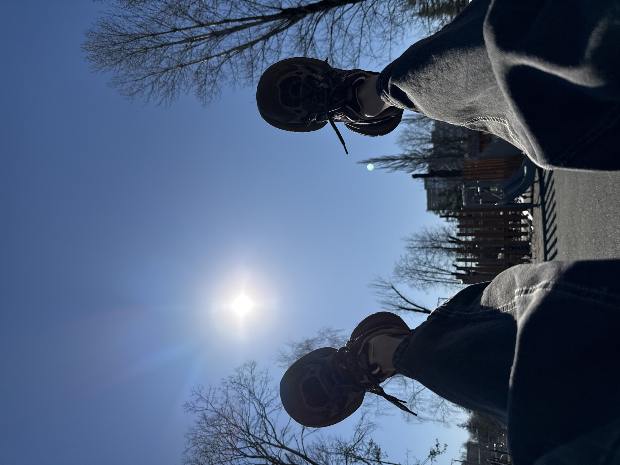 A low-angle photograph looking up towards the sky. The sun is visible in the sky, partially obscured by trees. In the foreground, the viewer sees two legs and feet, wearing dark pants and brown shoes. In the background, indistinct structures, likely buildings and playground elements, are present, hinting at an urban park location. Given the specified location of Rusanovo, Russia, it can be inferred that the photograph was taken there. The scene appears to be outdoors on a sunny day.