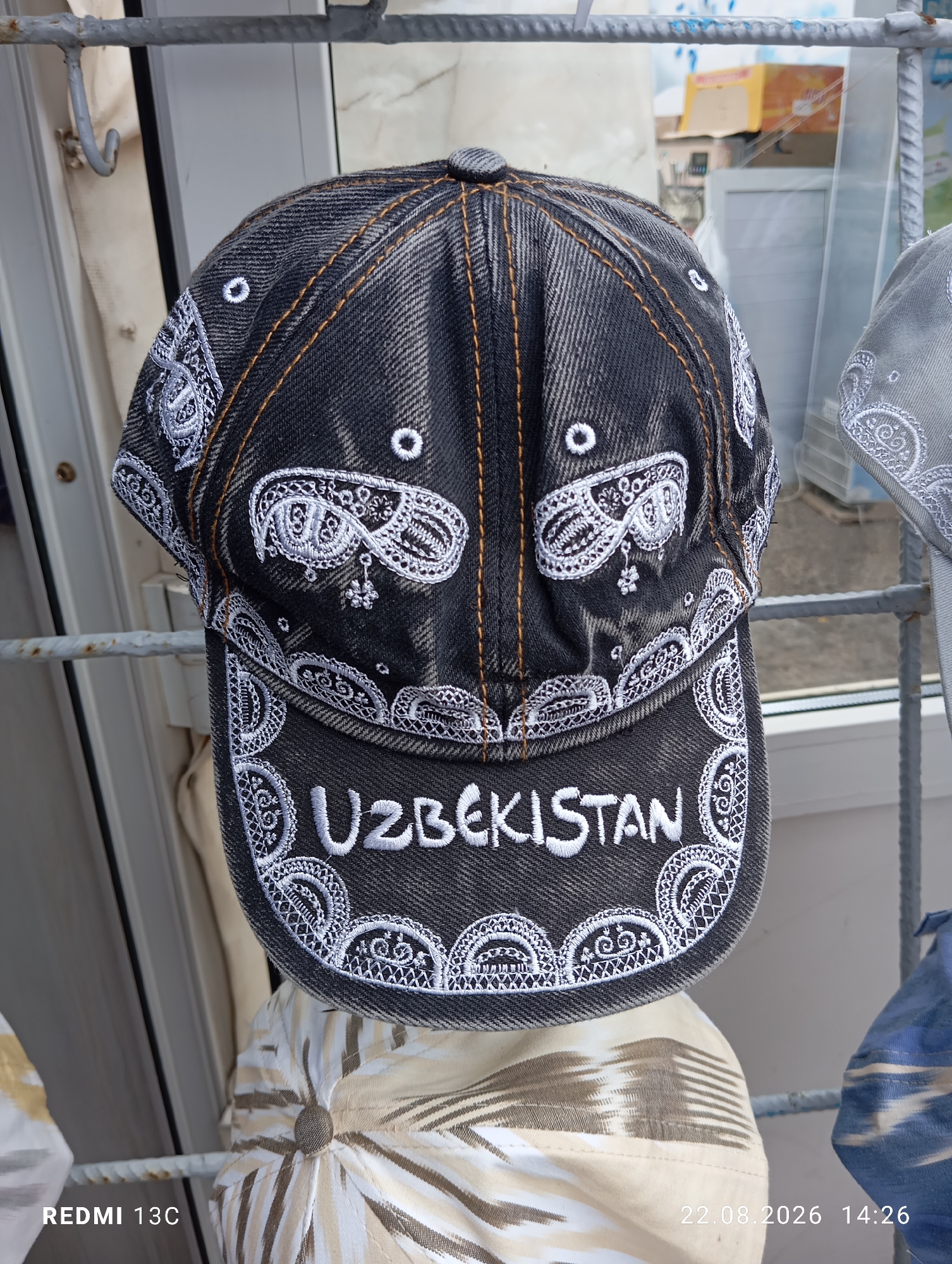 A close-up, eye-level shot shows a dark gray denim baseball cap with white embroidery. The embroidery features paisley-like patterns around the brim and the word "UZBEKISTAN" in white capital letters across the front. The cap is displayed outdoors, likely in a market or shop, with a metal grate and a window in the background. In the background, there is also a partially visible light-colored baseball cap with a patterned design. The bottom of the image includes a watermark that reads "REDMI 13C" and a timestamp "22.08.2026 14:26".