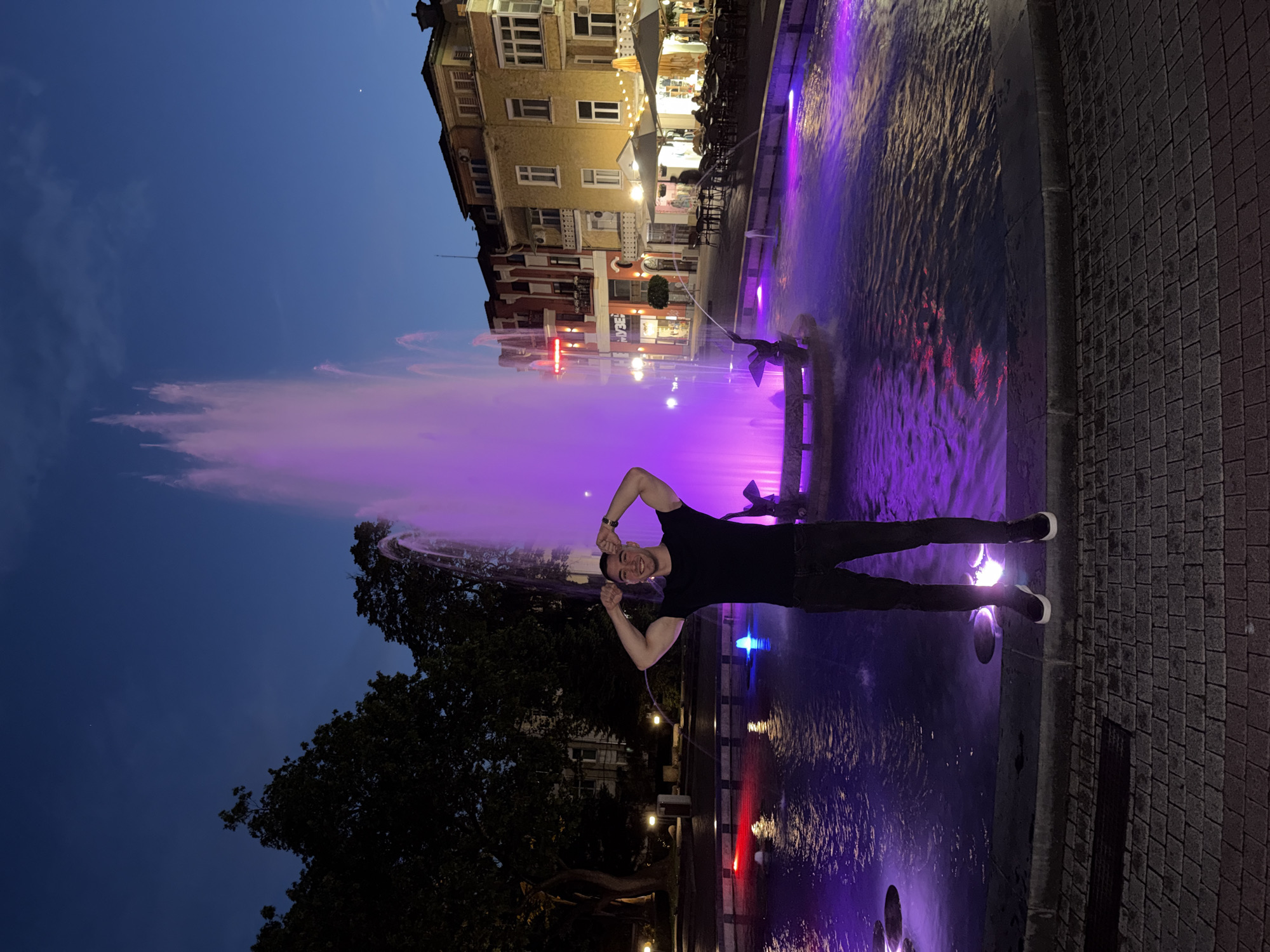 A medium shot captures a man standing in front of a fountain at night in Unknown, Unknown. The man is dressed in black pants, a black t-shirt, and shoes with white details. He has his arms raised, forming a heart shape with his hands above his head, and a smile on his face.
The fountain is illuminated with vibrant purple lighting, and the water jets create a luminous display. In the background, a multi-story building is visible, with lit windows and an outdoor seating area. The sky is a deep blue, suggesting dusk or early evening. The pavement around the fountain is made of gray bricks. A tree is visible to the left of the fountain. The lighting and composition suggest a lively urban environment.
