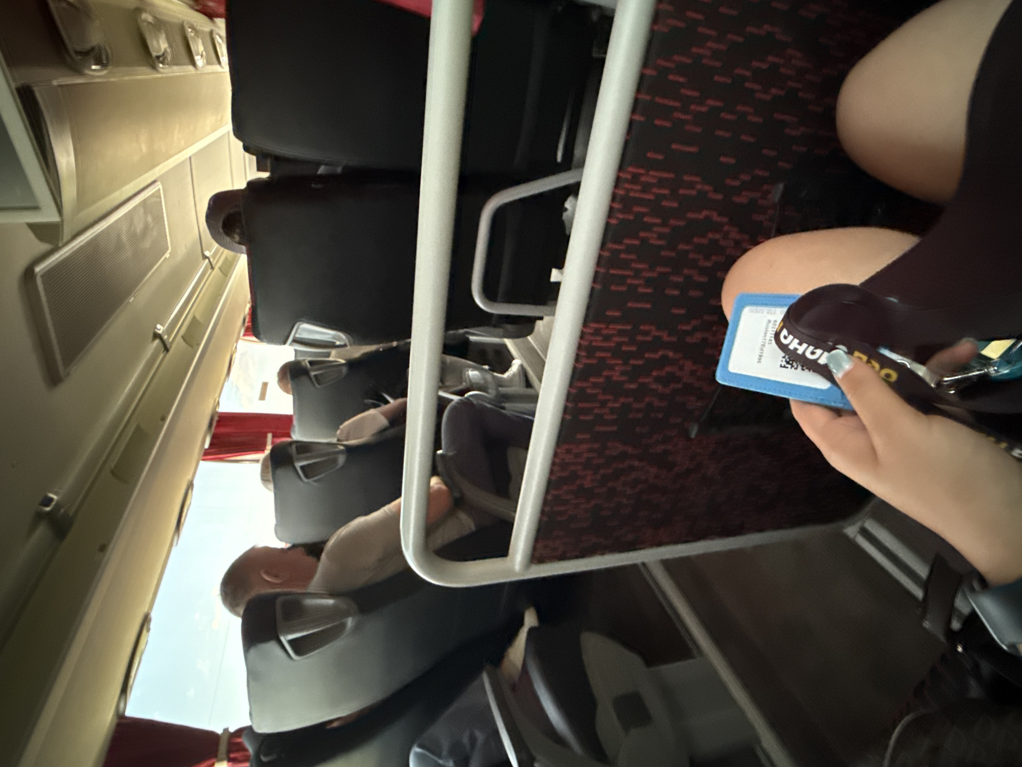 Inside a train car, passengers are seated in rows. Overhead, the train car's interior is visible. A passenger is holding a travel pass, and the background features patterned seat fabric. The destination on the pass is in Prague.