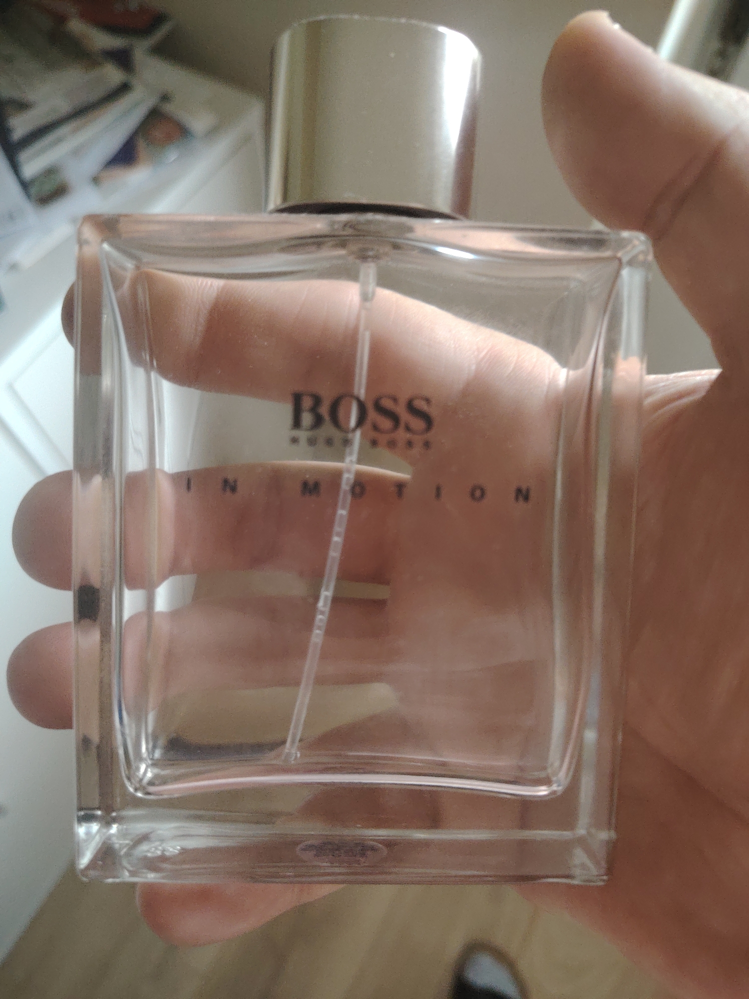 A hand is holding a clear glass perfume bottle, with the brand name "BOSS" and the text "IN MOTION" visible on the front. The bottle is square-shaped with a silver cap. The background is out of focus and appears to be an indoor setting with shelves and items on them. The image does not provide any information about the location in the city of Huntingdonshire, United Kingdom.