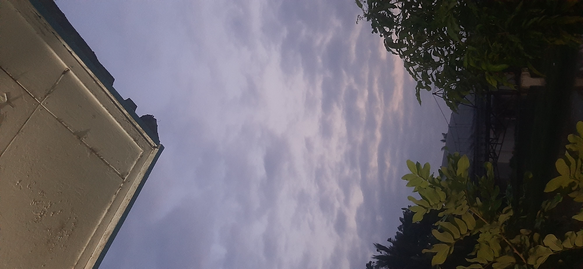 The image captures a view from below an overhang, looking up at a cloudy sky. The overhang appears to be part of a building or structure, with peeling paint visible on its surface. Below the sky, silhouetted trees frame the lower portion of the image. In the mid-ground, a structure resembling a bridge or overpass is visible, partially obscured by the trees and the dim lighting. The overall impression is of an overcast day in an urban or semi-urban environment, possibly during twilight. Without specific landmarks or contextual information, pinpointing the exact location within Jambi, Indonesia, is not possible from the visual cues alone. The scene is devoid of people.