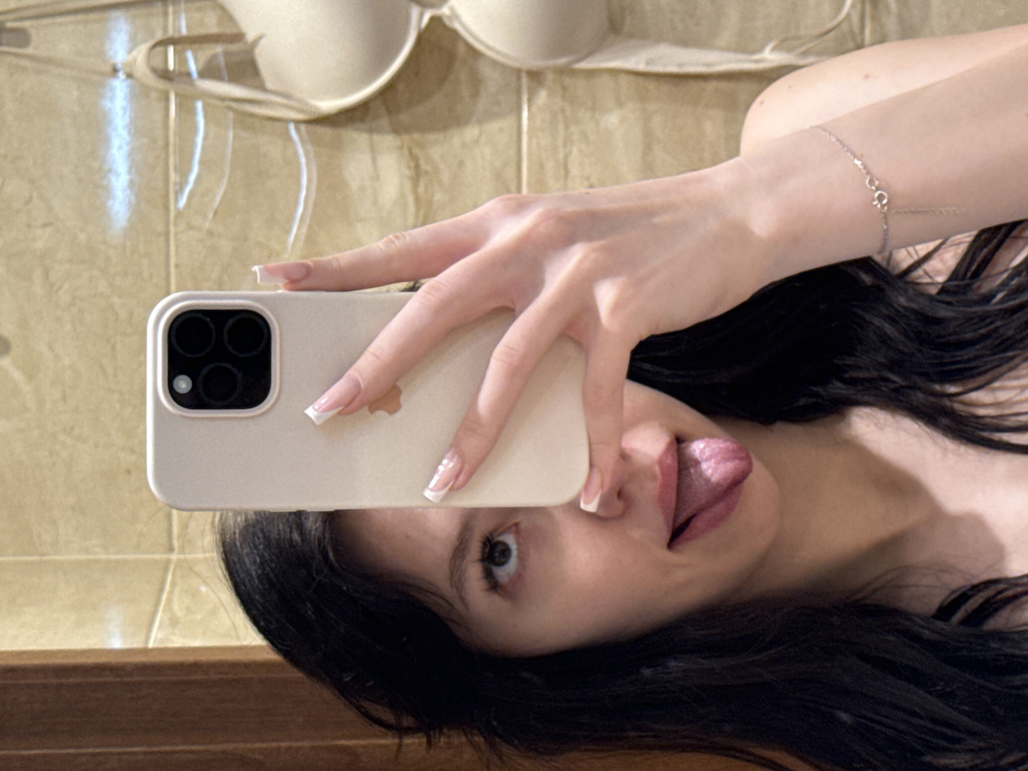 In a bathroom, a young woman with long, dark hair is taking a selfie in a mirror. She is holding a light-colored smartphone in her right hand, obscuring part of her face. Her tongue is sticking out, and she is looking upwards. A bra with beige cups is visible hanging on the wall behind her. The wall is tiled with a light brown, textured pattern. The scene appears to be indoors, likely in a residential setting. The location in Khimki, Russia, is not evident from the visual information alone.