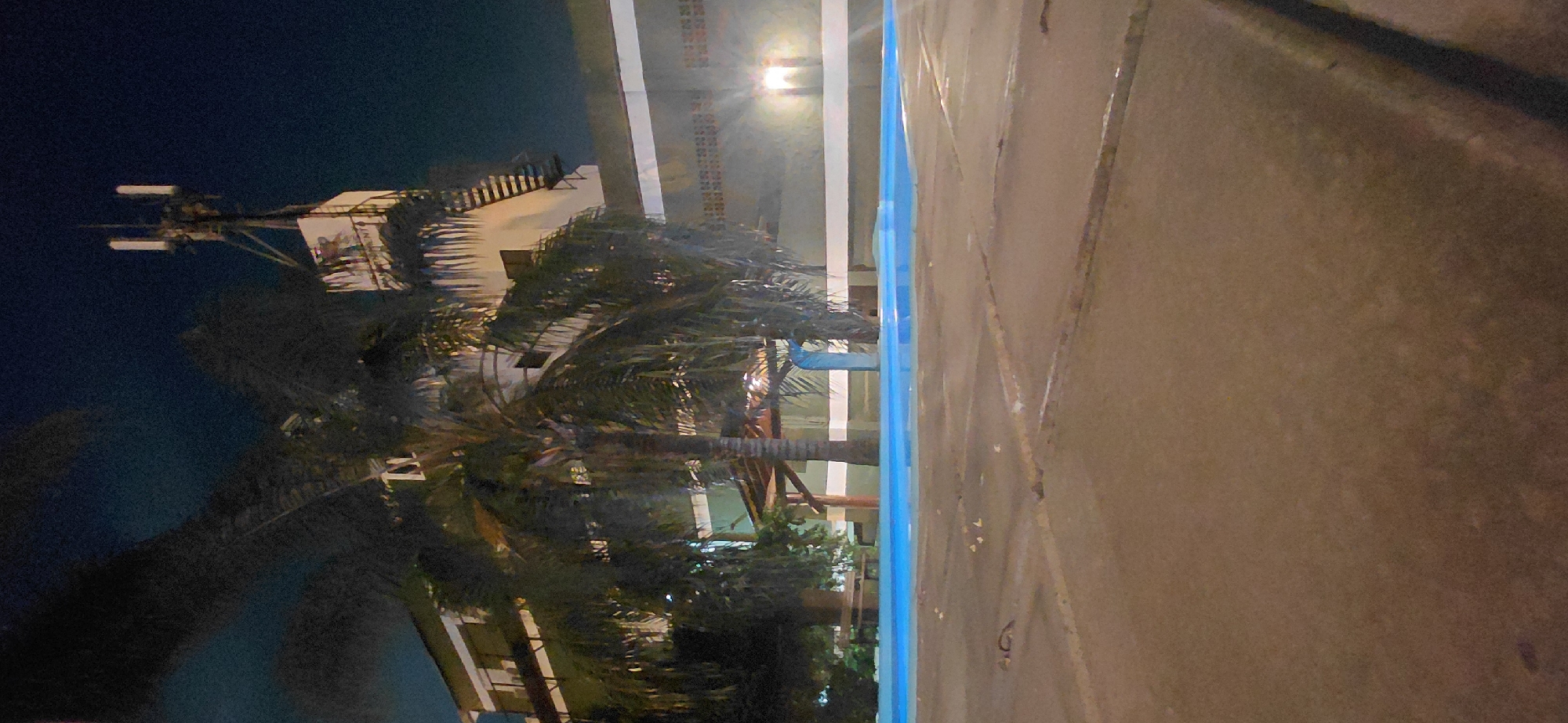 The image depicts a nighttime scene at an outdoor swimming pool area in Presidente Franco, Paraguay. A brightly lit, rectangular swimming pool with a vibrant blue surface is visible in the foreground. The pool is surrounded by a paved area with visible expansion joints. Beyond the pool, a multi-story building with white and green accents stands. On top of this building, a prominent telecommunications tower with multiple antennas is erected, illuminated against the dark sky. Palm trees with lush foliage are scattered throughout the scene, casting shadows and adding to the tropical ambiance. A rustic wooden structure, possibly a gazebo or a covered seating area, is situated near the pool. Exterior lighting illuminates parts of the building and the surrounding area, creating a stark contrast with the night sky. The overall environment suggests a recreational or lodging facility.
