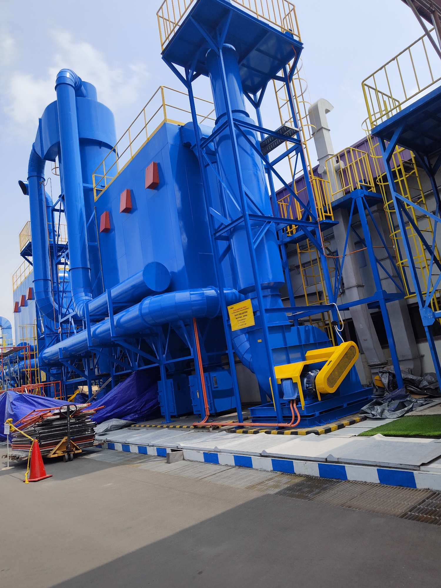 The image shows a large, bright blue industrial dust collector unit with yellow safety railings and platforms. The unit is situated outdoors on a paved surface with blue and white striped borders. Construction materials, including scaffolding and tarps, are visible around the base of the unit, suggesting an ongoing project. A red traffic cone is also present near a pile of stacked pipes. The sky is partly cloudy. The scene is located in Raci, Indonesia, a detail derived from the yellow warning sign attached to the dust collector which states "PT. LIMPIAN SUMBER SEJAHTERA" and "DUST COLLECTOR BUFFALO BPPO", indicating a company name and possibly a product model or location. The image captures a component of an industrial facility under construction or renovation. There are no people visible in the image.