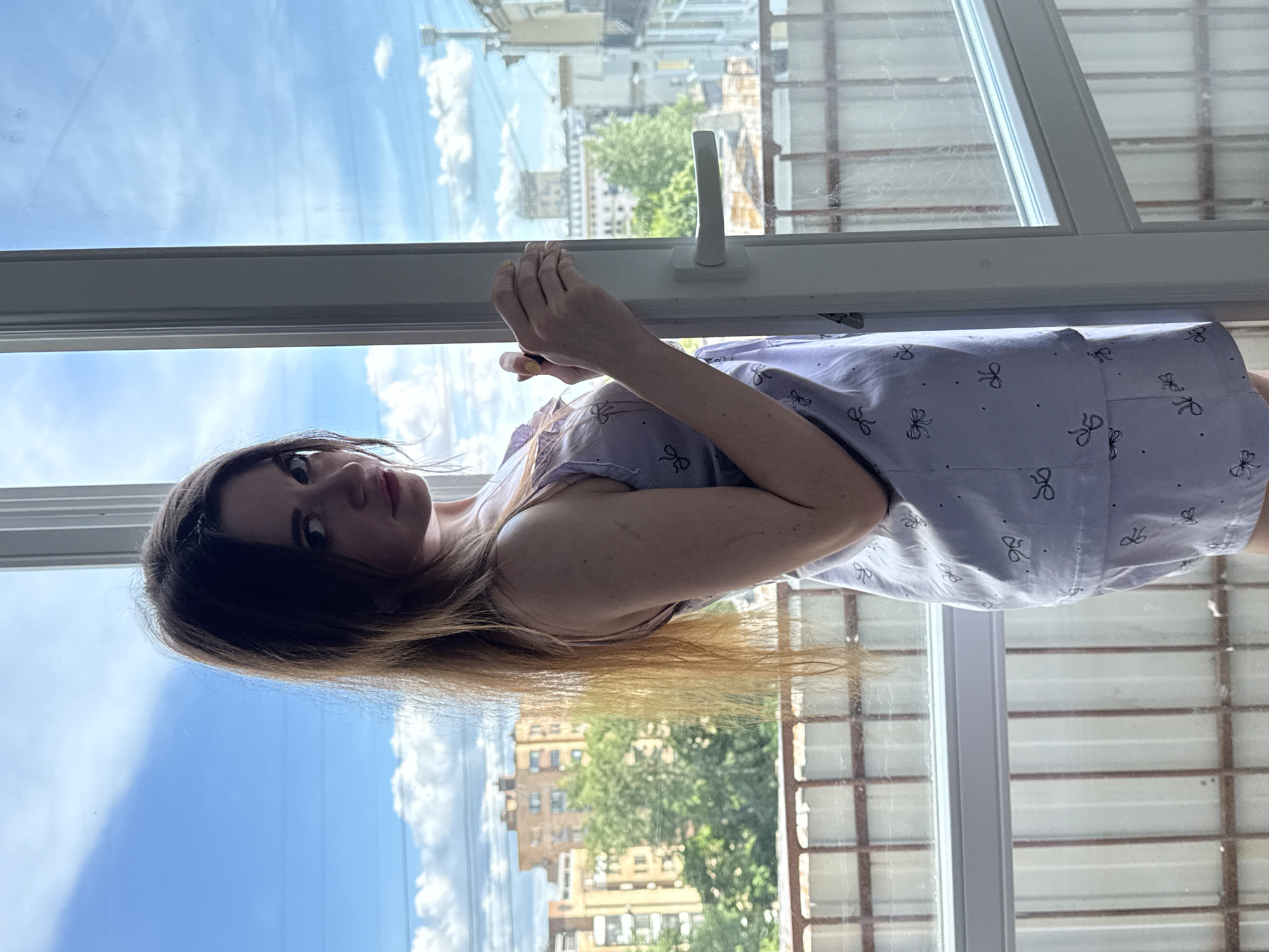 A woman with dark hair and blonde highlights is standing in front of an open window. She is wearing a light-colored dress with a bow pattern. The window provides a view of a cityscape, including buildings and trees, possibly within Moscow, Russia. The weather appears sunny with scattered clouds. The window is white with a visible handle. The woman is looking at the camera. Her hands are near the window frame.