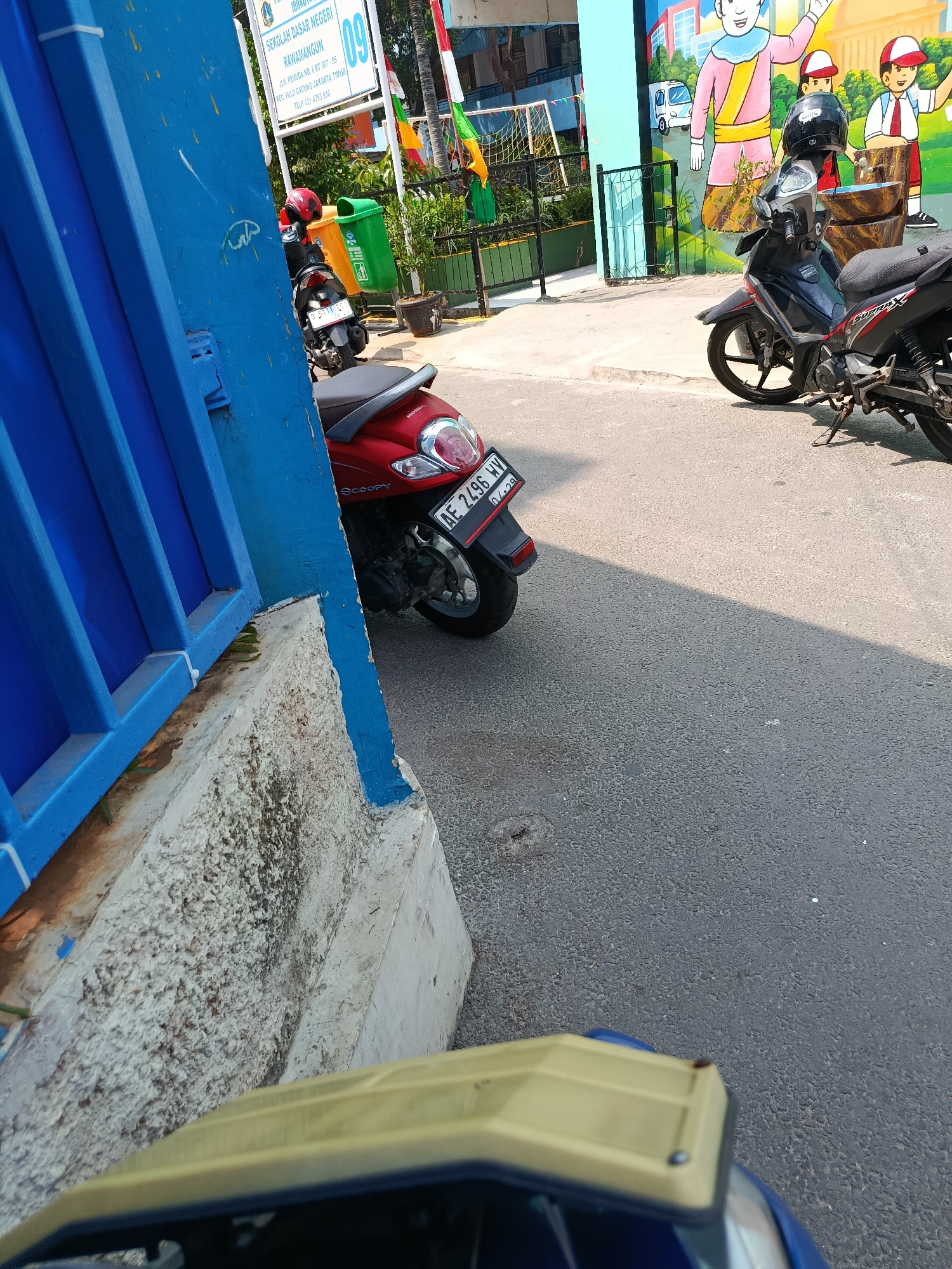 The image depicts a street scene in front of a school building, identified as "Sekolah Dasar Negeri Raharhamangcun" in East Jakarta, Indonesia. The foreground is dominated by a bright blue metal fence on the left. To the right of the fence, a red scooter with a license plate reading "AE 2496 HV" is parked, partially obscured by the fence. Further to the right, a black motorcycle with a helmet on its handlebars is also parked. Behind the motorcycles, a mural adorns the school building, featuring cartoon figures of children and adults, as well as a small white van. Two trash bins, one green and one yellow, are visible near the school entrance. The ground is paved with asphalt, and a shadow stretches across it, indicating a sunny day. The overall scene suggests a typical school environment during non-operational hours or a break.