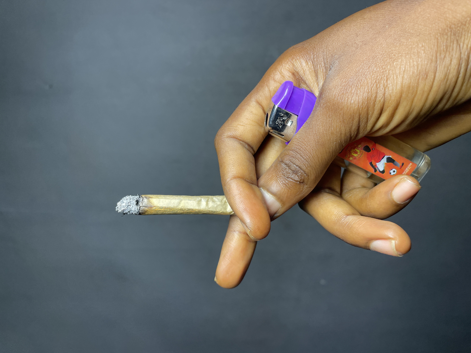 A close-up shot features a person's hand holding a lit, rolled cigarette or joint and two lighters against a plain dark gray background. The hand has a dark skin tone, with visible wrinkles and skin texture.
The rolled item, appearing to be a joint or hand-rolled cigarette made with light brown paper, is held between the index and middle fingers, extending towards the left. Its tip is heavily covered in white ash, indicating it has been burning for some time.
Two lighters are held in the palm and secured by the thumb and other fingers. One is a purple disposable lighter, with the metallic cap visible and the brand name "FAJ" partially etched into it. The second lighter, partially obscured by the hand, is clear plastic and contains a liquid, possibly lighter fluid. It features a sticker depicting a soccer player in a red jersey, white shorts, and black socks, kicking a soccer ball. Below the player, the word "MANCHESTER" is clearly visible, suggesting a Manchester United Football Club theme.
The lighting is soft and even, highlighting the details of the hand and objects. The background is uniformly dark, providing no additional environmental cues. The image captures a moment of smoking or preparing to smoke. The context for this scene is Nkpor, Nigeria.