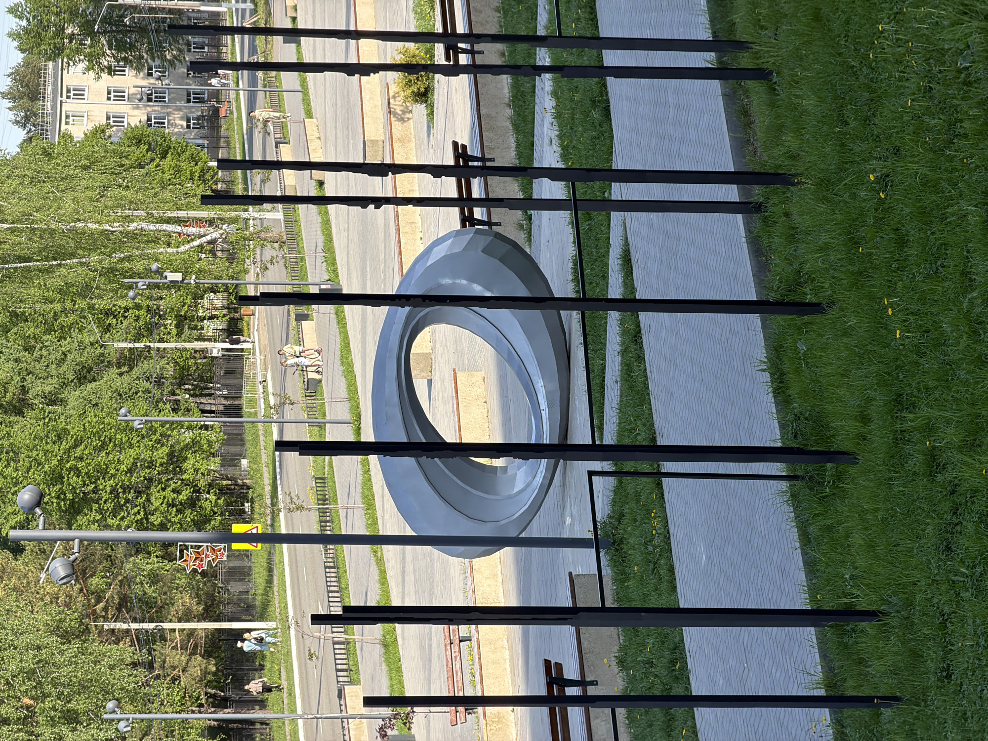 The image depicts an outdoor urban scene in Chaikovsky, Russia. A large, gray, donut-shaped sculpture is centrally located, framed by black vertical bars, likely part of a decorative fence or barrier. The ground surrounding the sculpture is a mix of gray paving stones and green grass. In the background, across a pedestrian walkway, are benches, trees, street lights, and a multi-story building. Several people are walking or riding bikes along the walkway. The scene is well-lit, indicating it is daytime.