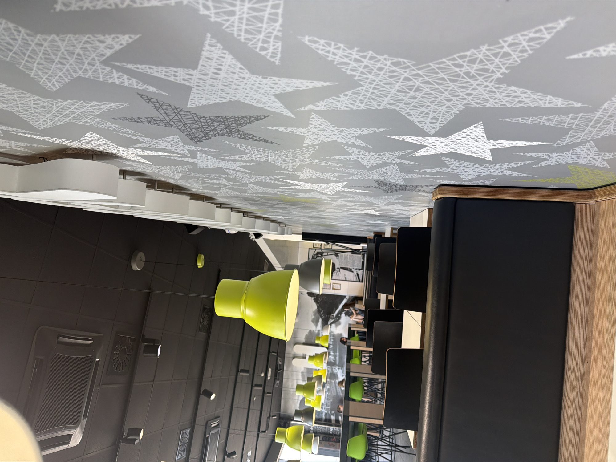 The image captures an interior scene, likely a cafe or restaurant, with a strong emphasis on the ceiling design and seating areas. The ceiling is a light gray with a repeating pattern of white and dark gray stars, some outlined and some filled with a crisscross texture. White, curved light fixtures hang from the ceiling.
Below the ceiling, rows of dark gray seating booths are visible, separated by wooden partitions. A bright green pendant lamp hangs prominently in the foreground, and several other green and gray pendant lamps are seen further back. Some of these lamps appear to be the same style as the one in the foreground.
In the background, through the seating area, glimpses of other patrons can be seen sitting at tables, some in green chairs. The overall impression is a modern, stylish indoor space, possibly during daylight hours, though the lighting is artificial. There is no visible text in the image. The location context provided is Kyiv, Ukraine.
