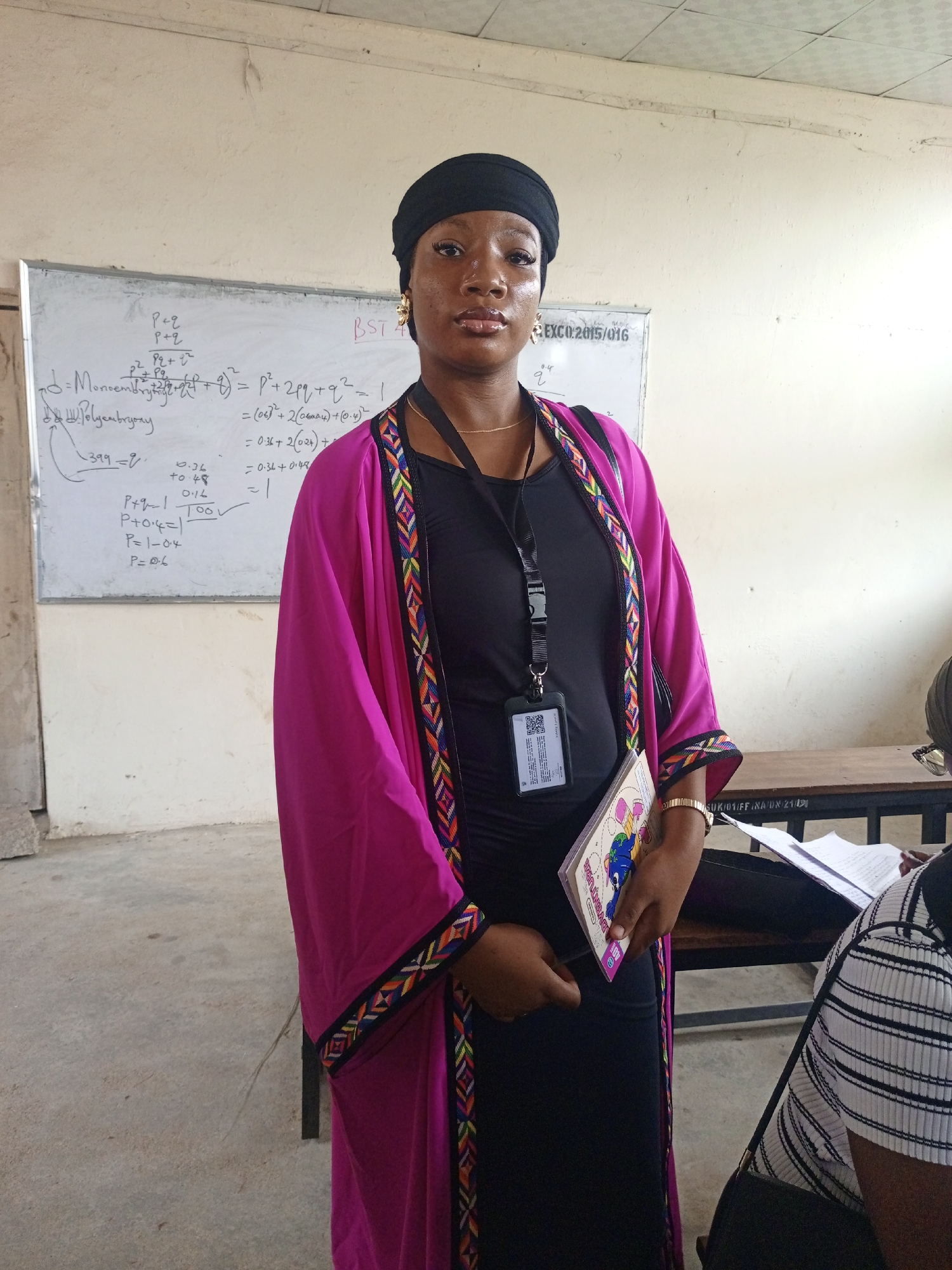 A young woman stands in what appears to be a classroom in Keffi, Nigeria, looking directly at the camera. She wears a black head wrap, a black top or dress, and a vibrant magenta/fuchsia open cardigan adorned with a colorful geometric trim along the edges. A thin gold chain is visible around her neck, and a gold-colored bracelet is on her left wrist. Around her neck hangs a black lanyard with an ID badge holder, which shows a QR code and some text. She holds a small book with "ADVENTURE" visible on its cover.
Behind her, a white board is filled with handwritten equations and notes, suggesting an academic or scientific context. Visible text includes "EXCO.2015/016", "BST 4", "P+Q", "P²+2pq+q²=1", "Monocembryony", and "Polyembryony", along with mathematical calculations like "(0.6)²+2(0.24)+(0.4)²", "0.36+0.48", and "P=0.6". To her right, part of another person, wearing a black and white striped shirt, is visible, seated at a wooden desk or bench with papers on it. The desk itself has a partially visible label: "SUK/01/FF.NA.UN.21/". The room has a white wall, a concrete floor, and a tiled ceiling with visible wiring. The lighting suggests it is daytime. The woman maintains a neutral and composed expression.