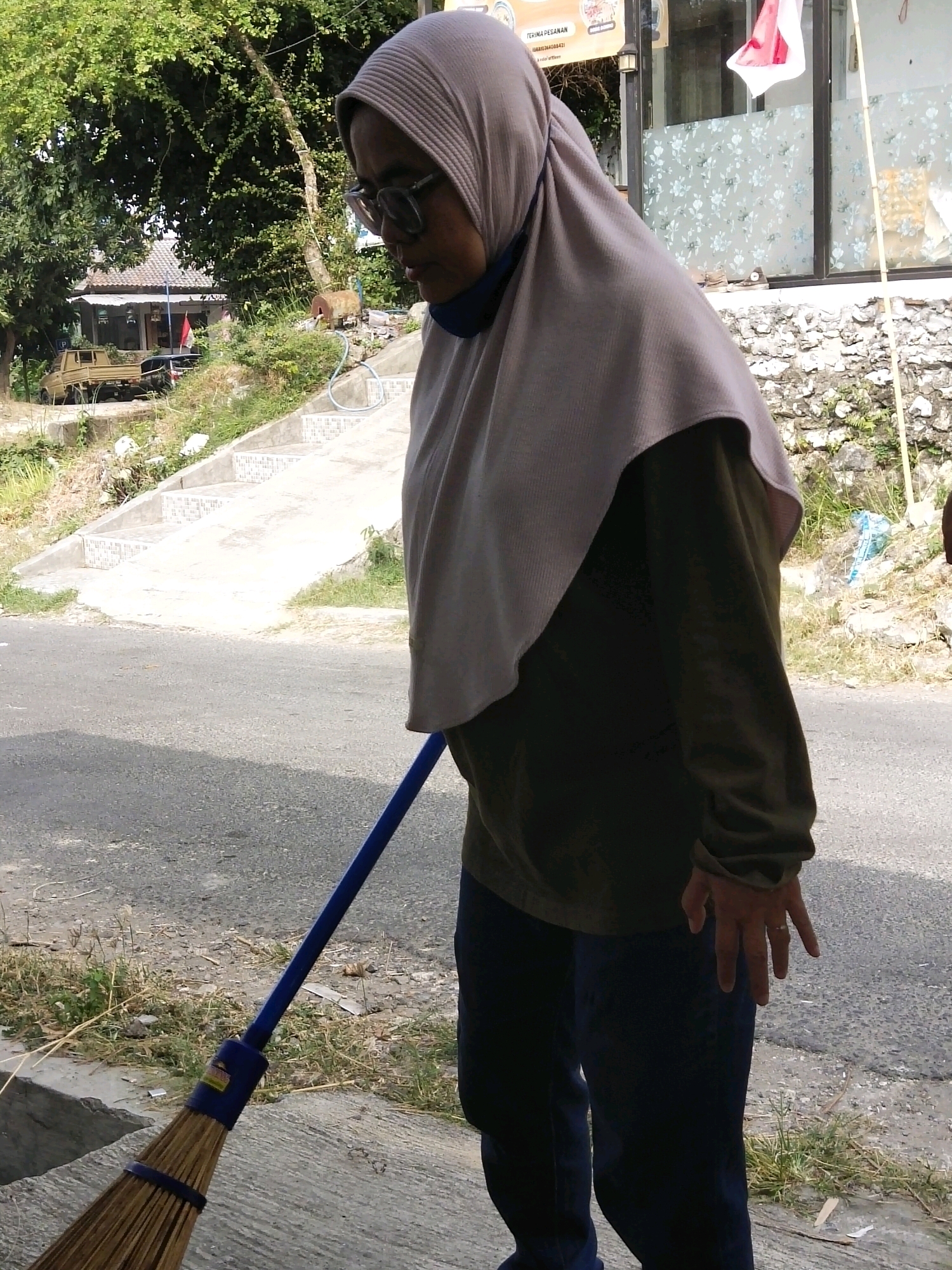 An Indonesian woman wearing a light purple hijab and glasses is sweeping the ground with a broom. She is dressed in a dark olive-green long-sleeved shirt and dark blue pants. The scene appears to be outdoors, near a road, with a stone wall and some vegetation in the background. There are concrete steps leading up a slope to a building with a red and white flag displayed. A small truck is parked in front of the building. The image has been captured from a medium distance, focusing on the woman and her activity.
