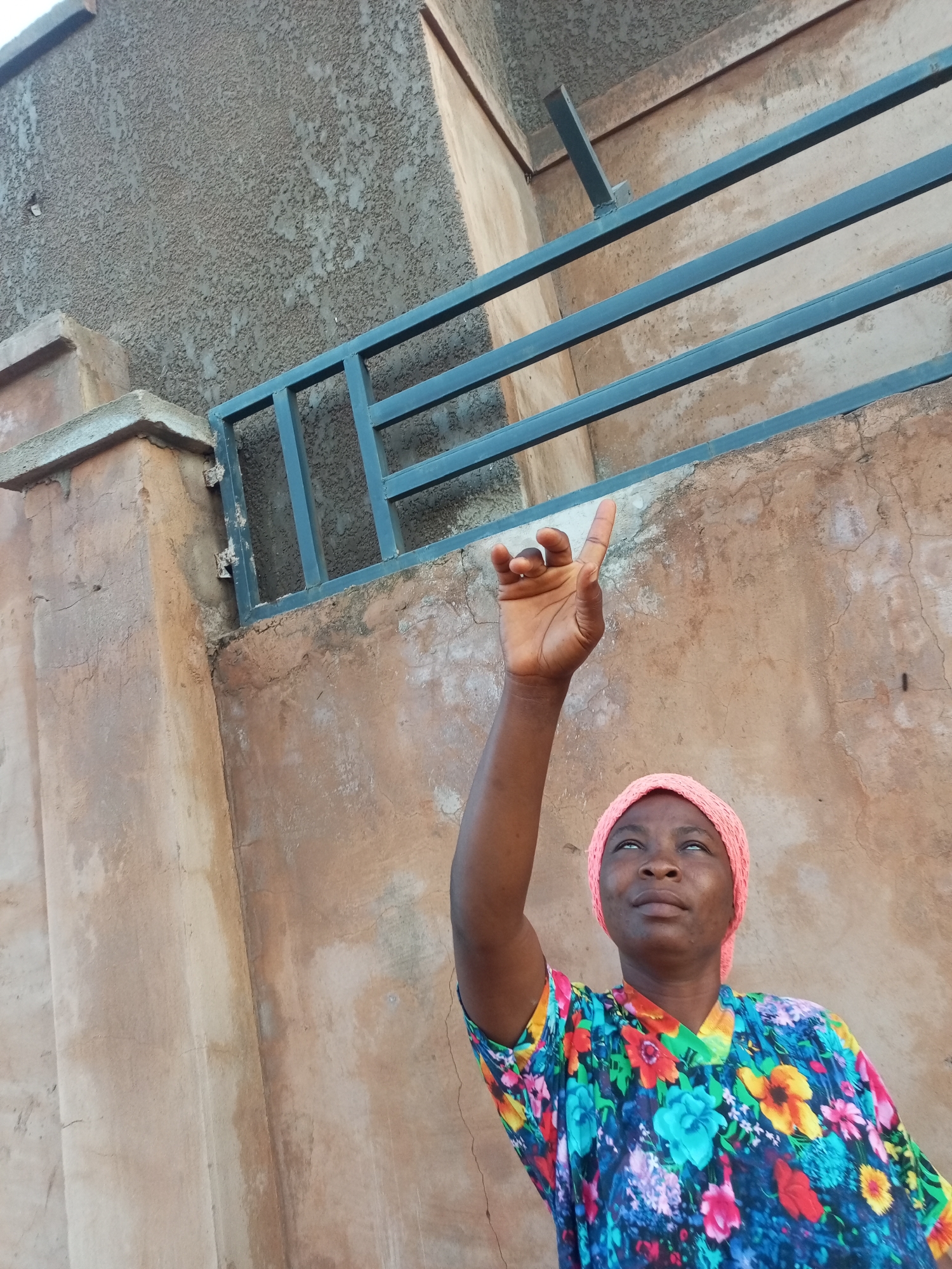 A woman wearing a pink headscarf and a colorful floral print shirt is looking up and pointing her index finger towards a metal railing. She is standing in front of a textured wall and a concrete structure. The scene appears to be outdoors, possibly in an urban environment. The lighting suggests it is daytime. The background shows a concrete wall with a rough texture and some patches of discoloration. There is a dark blue metal railing installed on top of the wall.