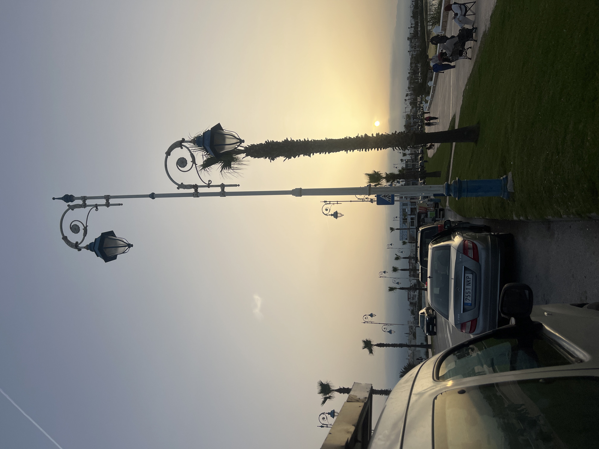The image captures a sunset scene along a coastal promenade in Martil, Morocco. The sun, a bright golden orb, is low on the horizon, casting a warm, luminous glow across the sky, which transitions from yellow and orange near the sun to a lighter blue-grey overhead.
In the foreground and midground, ornate street lamps with dark blue and white scrolling metalwork and classic lantern designs stand tall, some silhouetted against the bright sky. Several palm trees with tall, slender trunks and visible fronds are also prominent, especially a large one near the center, appearing almost completely silhouetted by the sun.
Along a paved area next to a strip of green grass, several people are visible in the distance. Some are sitting on benches, while others appear to be strolling, their figures largely silhouetted. Further back, the land meets the hazy horizon.
On the right side of the image, a row of parked cars lines a road. The closest vehicle in the foreground shows part of its roof and window. Directly behind it, a silver Mercedes-Benz C200 is visible from the rear, displaying the license plate "7551 NKP" and the "C 200" model designation. Other cars are parked further down the road. A blue circular road sign with a white arrow is partially visible on a pole in the midground. The overall atmosphere is peaceful, capturing the end of the day.