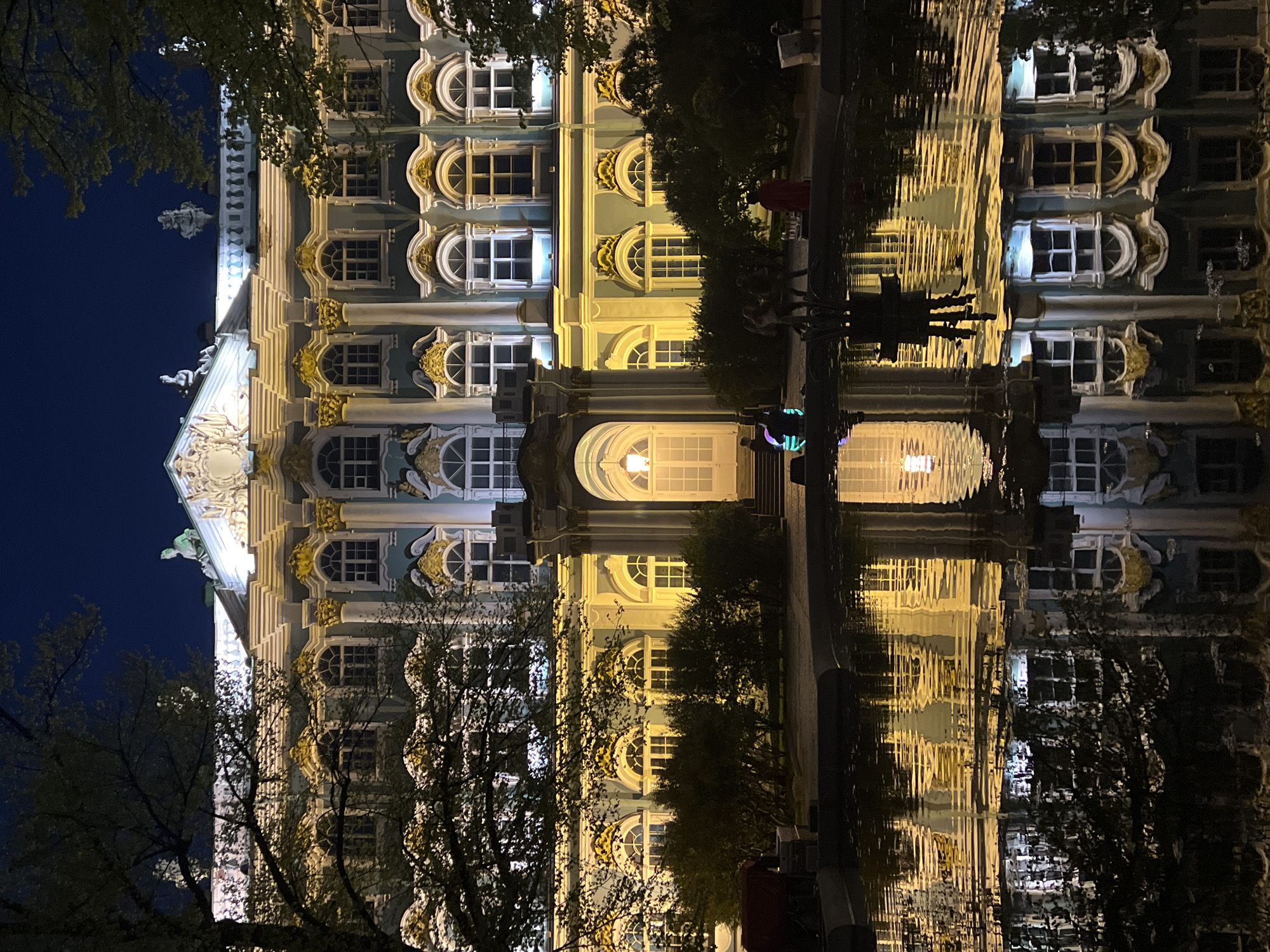 The image depicts the Winter Palace in Saint Petersburg, Russia, at night, as reflected in a body of water. The building is elaborately designed, featuring numerous windows with arched tops, decorative ornamentation, and sculptures on the roofline. The structure is illuminated, casting a warm yellow glow against the dark blue night sky. Trees are visible in the foreground, framing the building on both sides. In the water's reflection, indistinct figures of people can be seen along the waterfront. The water surface is slightly rippled, distorting the reflected image of the palace.
