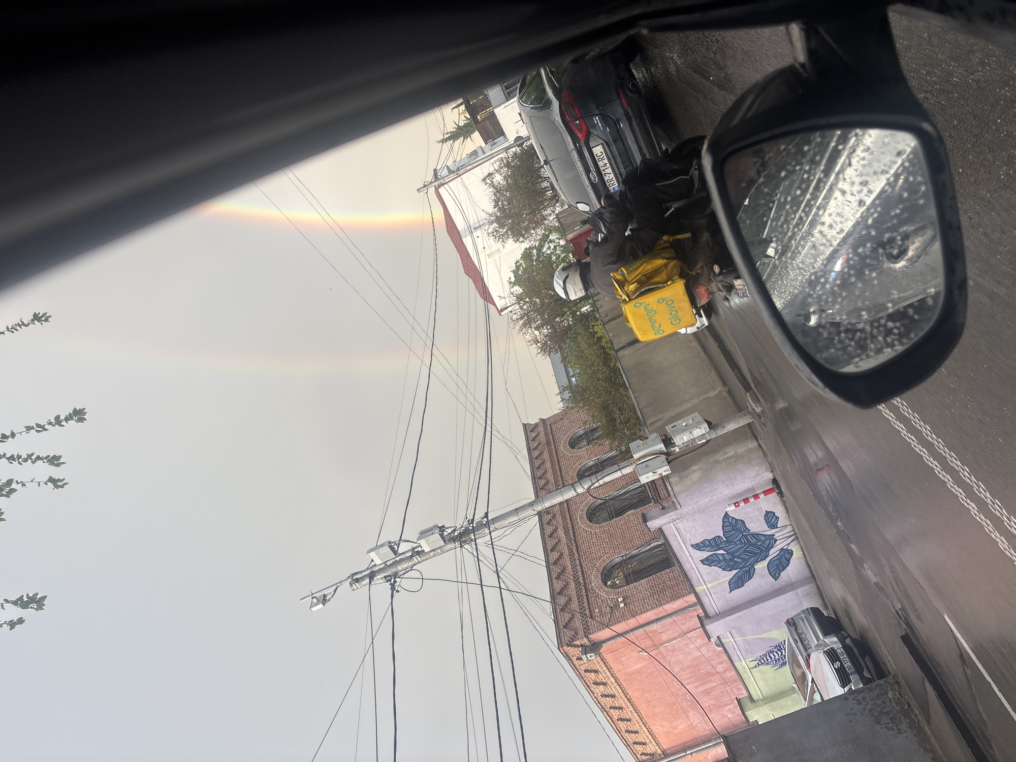 The image, taken from inside a vehicle, shows a rainy street scene with a rainbow visible in the sky. A motorcyclist with a Glovo delivery bag is next to a parked car. A building with brick sections and arched windows stands to the right, with a mural of leaves painted on the wall next to it. Utility poles and wires are visible overhead. The scene appears to be in Tbilisi, Georgia, based on the license plate "RR-714-RC" visible on the parked car. The side mirror of the vehicle is visible, covered in raindrops.