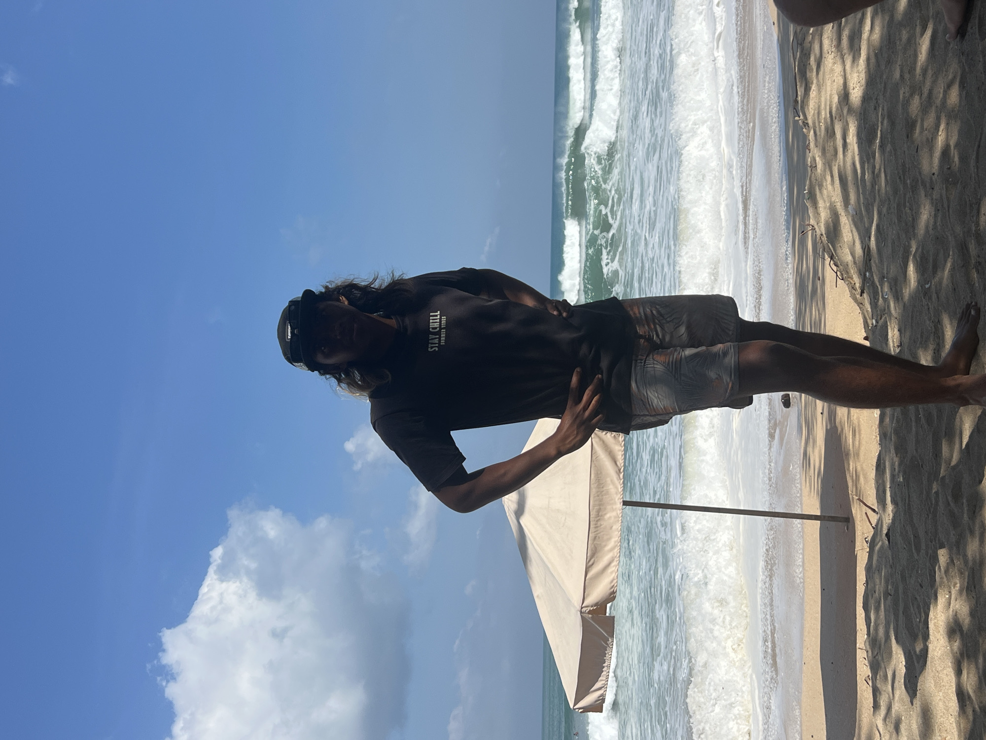 A person stands on a sandy beach in Kuta, Indonesia, facing the camera. They are wearing a dark t-shirt with the text "STAY CHILL" and "SUMMER VIBES" visible, along with patterned shorts and a black cap. Their hands are placed on their hips. Behind them, the ocean displays waves with white crests, and a beige beach umbrella stands to their left. The sky is a clear blue with scattered white clouds. The sand is textured with shadows cast from overhead foliage.