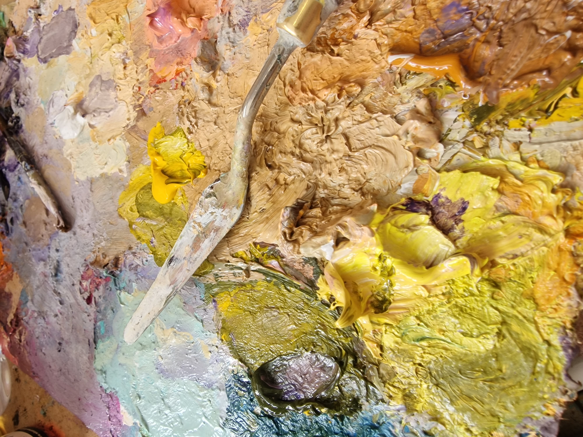 A wooden artist's palette is depicted at a high angle, centered in the frame, covered with thick, mixed oil paints. This scene is located within an artist's studio or workspace in Veliko Tarnovo, Bulgaria. The palette features a wide array of colors, including prominent areas of bright yellow, various shades of olive and dark green, light blue, beige, earthy brown, orange-pink, purple, white, and grey. Paint textures range from smooth, freshly squeezed dollops, such as a distinct bright yellow accumulation, to heavily impastoed, swirled, and blended sections indicating mixing. A silver-handled metal palette knife rests diagonally across the upper-middle of the palette, its blade coated with paint residue. Faint impressions suggestive of paintbrushes are discernible in the blurred background. No individuals are visible in the frame. The scene conveys evidence of recent or ongoing artistic creation.