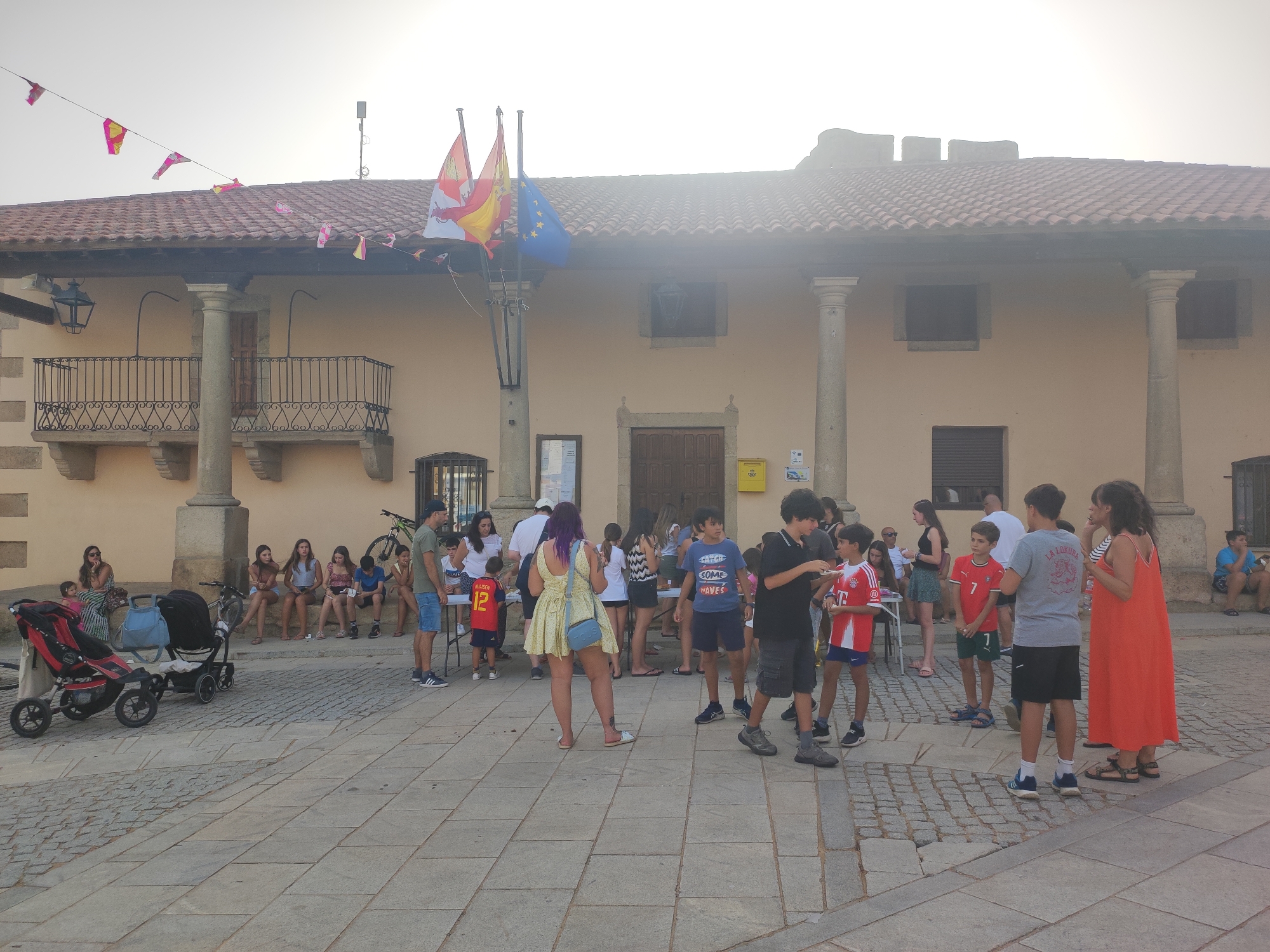 The image depicts a sunny day in the city of Sobradillo, Spain, likely during a local event or festival, given the presence of flags and a gathering of people. The scene is set in a paved plaza in front of a light-colored building with a red-tiled roof and multiple arched windows, some with decorative balconies. Two large columns flank the entrance of the building.
In the foreground, a group of people are milling about. Several children and young adults are visible, some are standing and conversing, while others appear to be observing something or waiting. There is a red and black stroller on the left side of the image, suggesting the presence of young families.
Several flags are displayed prominently on poles in front of the building. These include what appears to be the flag of Spain, and a European Union flag, along with smaller decorative flags strung across the building.
The atmosphere seems relaxed and casual. The people are dressed in summer clothing, and the lighting suggests it is daytime. The cobblestone pavement and the architectural style of the building point to a historic or traditional setting within the town.