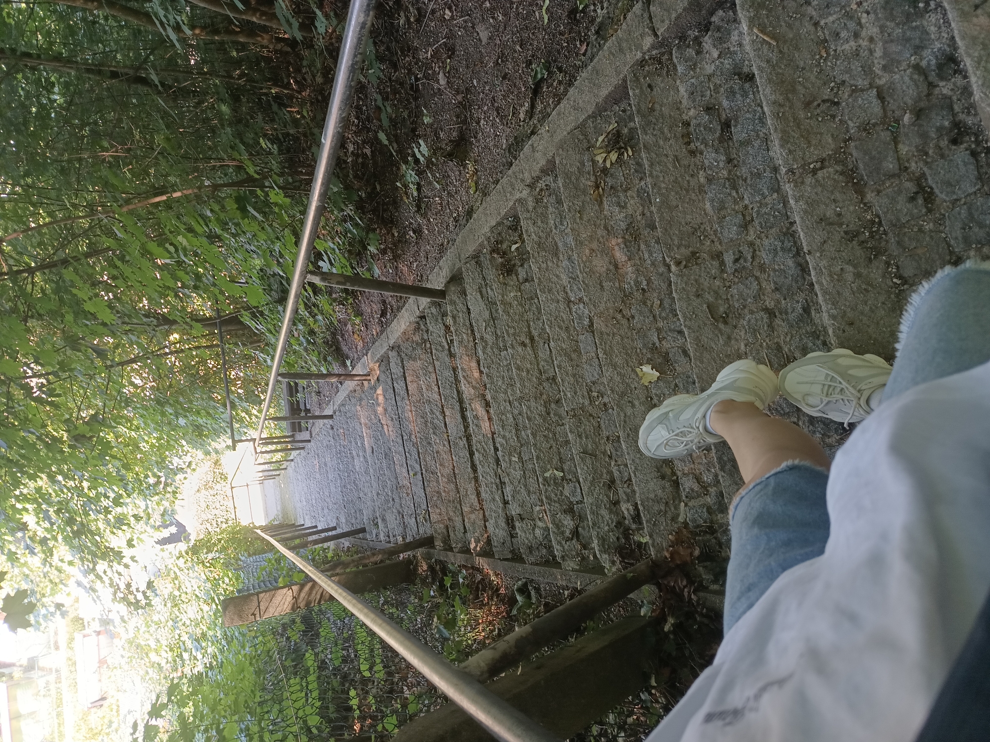 The image shows a person wearing white sneakers and denim shorts standing on a stone staircase descending into a wooded area. A metal handrail runs along the right side of the stairs. The staircase is bordered by a chain-link fence on the left, with lush green trees and foliage on both sides. The sun is shining through the trees, creating dappled light on the stairs and the surrounding environment. The scene suggests an outdoor location, possibly a park or a hiking trail. The city of Neuötting, Germany is not explicitly identifiable from the visual cues in the image alone, as there are no specific landmarks or signs to indicate this location.