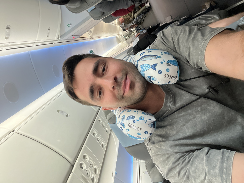 The scene depicts the interior of a commercial aircraft cabin. A male individual, appearing to be in his late 20s to early 30s, is centered in a self-portrait. He has short dark hair, light facial hair, and a slight smile, looking directly into the camera. He wears a grey short-sleeved t-shirt and a light blue and white neck pillow adorned with blue fish patterns and the word "GREECE" repeated multiple times. A black cord necklace with a small black pendant is visible around his neck. The aircraft cabin features white overhead compartments, passenger service units, and overhead lighting, including a blue accent light strip along the ceiling. Several other passengers are partially visible further down the aisle, seated in their respective seats. The context of the "GREECE" themed pillow and the specific prompt request places this travel event as relevant to the Paiania Municipal Unit, Greece, likely pertaining to Athens International Airport.