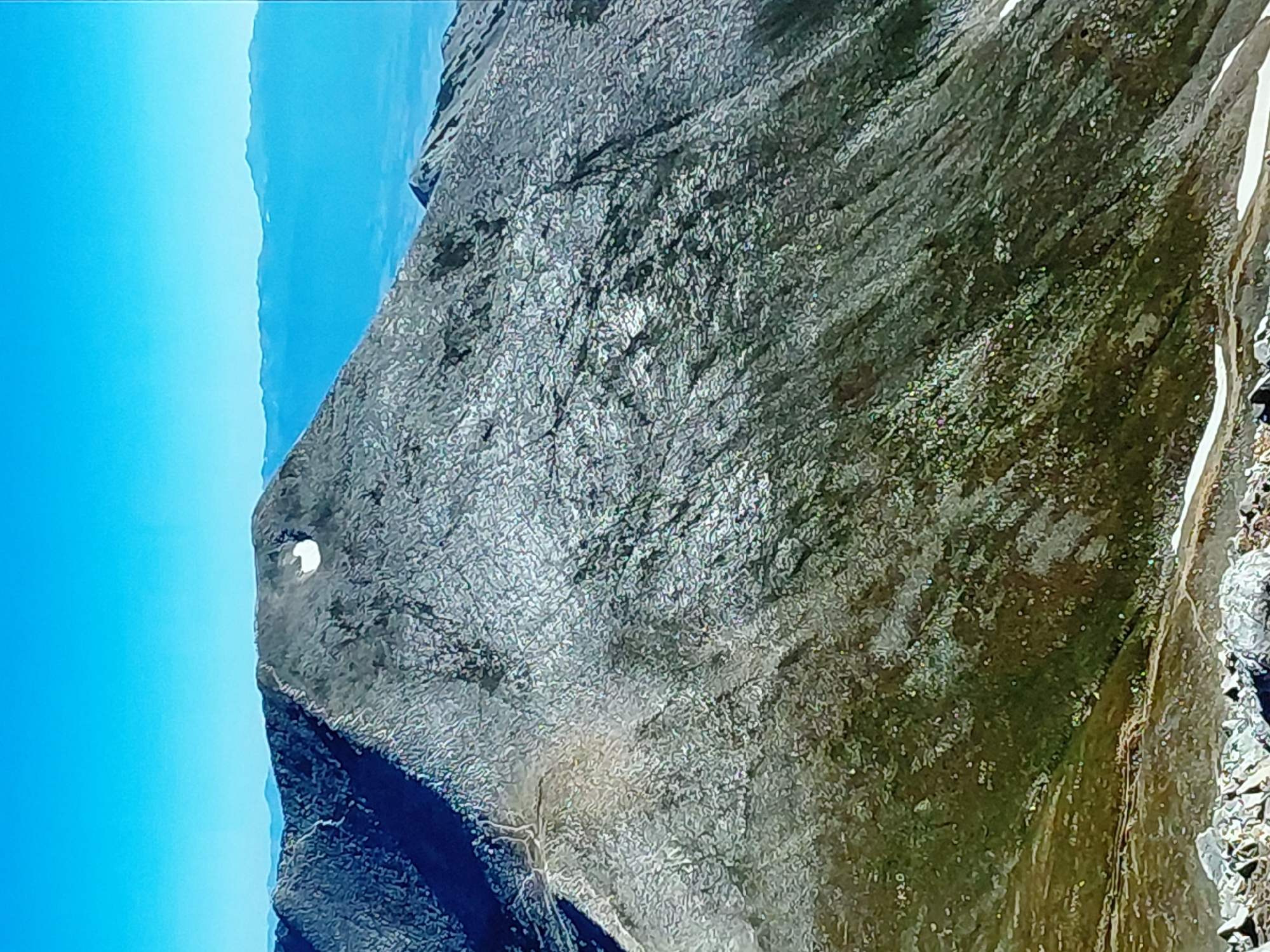 The image displays a rugged mountain landscape under a clear, bright blue sky, identified as being in the vicinity of Krumovo, Bulgaria. A large, grey mountain dominates the central and foreground, its steep slopes covered in a highly textured surface of countless small, light-colored rocks and scree, interspersed with darker rock formations. A prominent, roughly oval-shaped white patch, likely snow or ice, is visible near the summit on the right side of the main peak.
On the lower left and center-left slopes of the mountain, patches of dark green vegetation and brown earth are present, showing winding, faint lines that appear to be trails or erosion paths. The immediate foreground consists of scattered, angular grey boulders and rocks. Smaller patches of white snow are also visible in shaded areas on the lower right slope.
In the background, a long, undulating range of mountains or hills appears hazy and in lighter shades of blue due to atmospheric perspective, forming the horizon line beneath the expansive, clear cerulean sky. No individuals, animals, or man-made structures other than implied trails are visible.