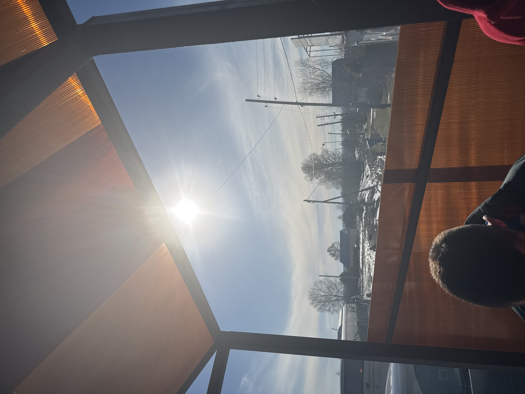 View through a framed window. The foreground shows the corner of a building or structure with orange siding and dark grey framework. The sun shines through the pane, creating a bright glare. The background is the exterior view of the city of Gory, Russia, with a blue sky, light clouds, and electric poles with lines. Buildings and trees line the street. A person's head is partially visible in the lower right corner, and part of their red clothing is in the upper right corner.