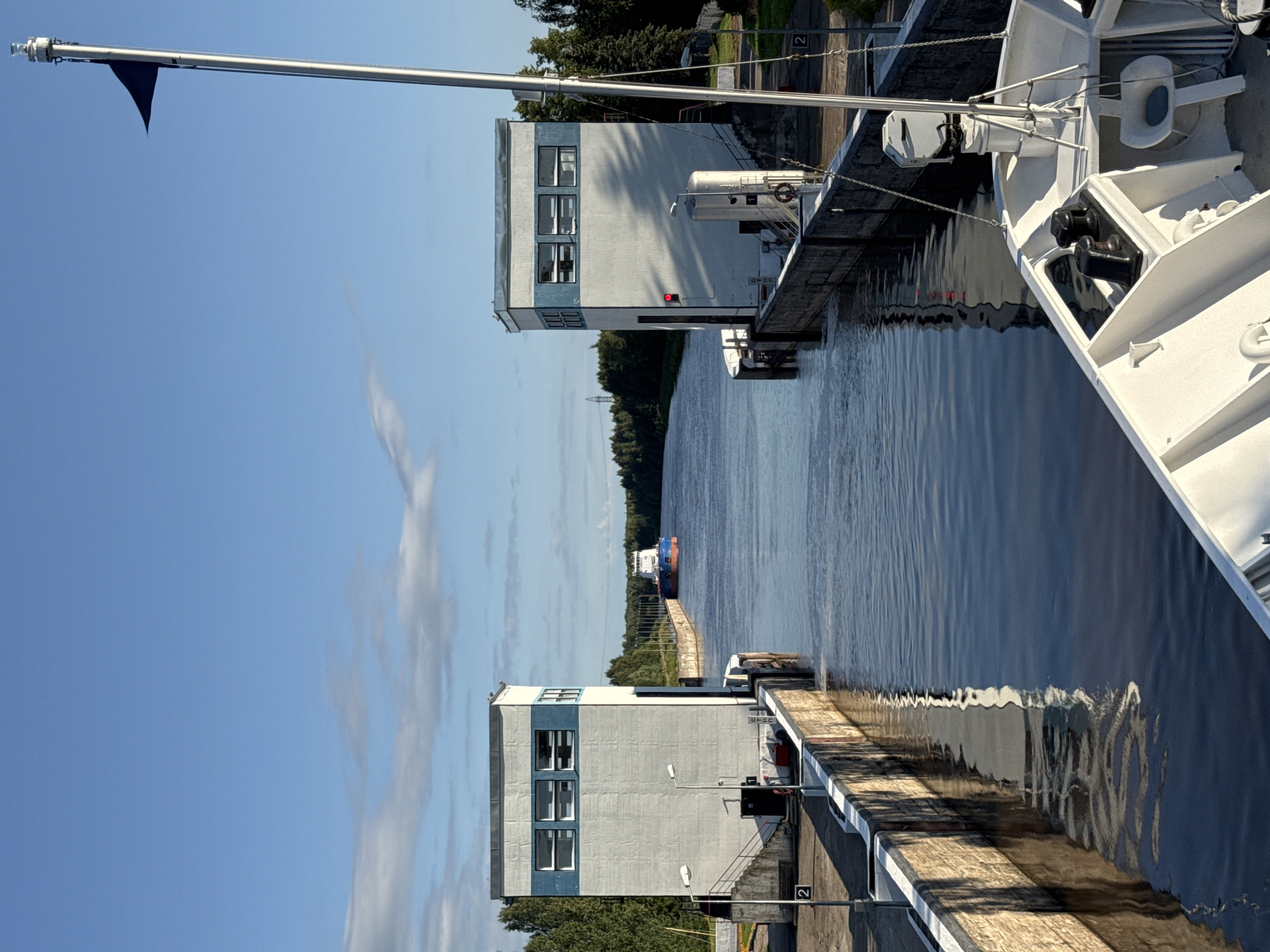The image depicts a view from the deck of a white vessel looking towards a canal lock in Vytegra, Russia. The lock is flanked by two identical concrete structures with blue-striped windows. In the distance, a large cargo ship, painted with a red and blue hull, is navigating the canal. The water is calm and reflects the surrounding structures and the clear blue sky with scattered white clouds. Trees line the banks of the canal in the distance. A metal flagpole with a dark flag is visible on the right side of the frame, extending into the sky. Essential details about people are not visible. The scene captures a moment of transit through the canal lock system.