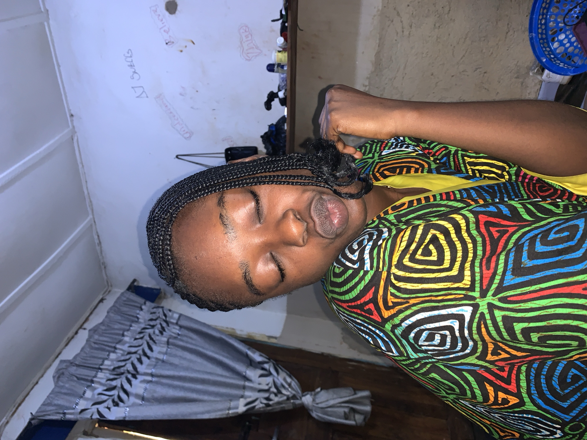 The image shows a close-up of a young Black woman with her eyes closed and her lips pursed, as if blowing a kiss. She has dark braided hair and is wearing a brightly colored garment with a geometric pattern in black, red, green, yellow, blue, and white. She appears to be indoors in a room with white walls. A gray curtain with a leaf pattern hangs to the left of the frame, and a shelf with various items, including bottles and a blue basket, is visible in the background. There is writing on the wall, including what appears to be "AJ Suells" and "Believe". The overall impression is a casual and personal moment captured in an informal setting, likely within a home in Rafin Par, Nigeria.