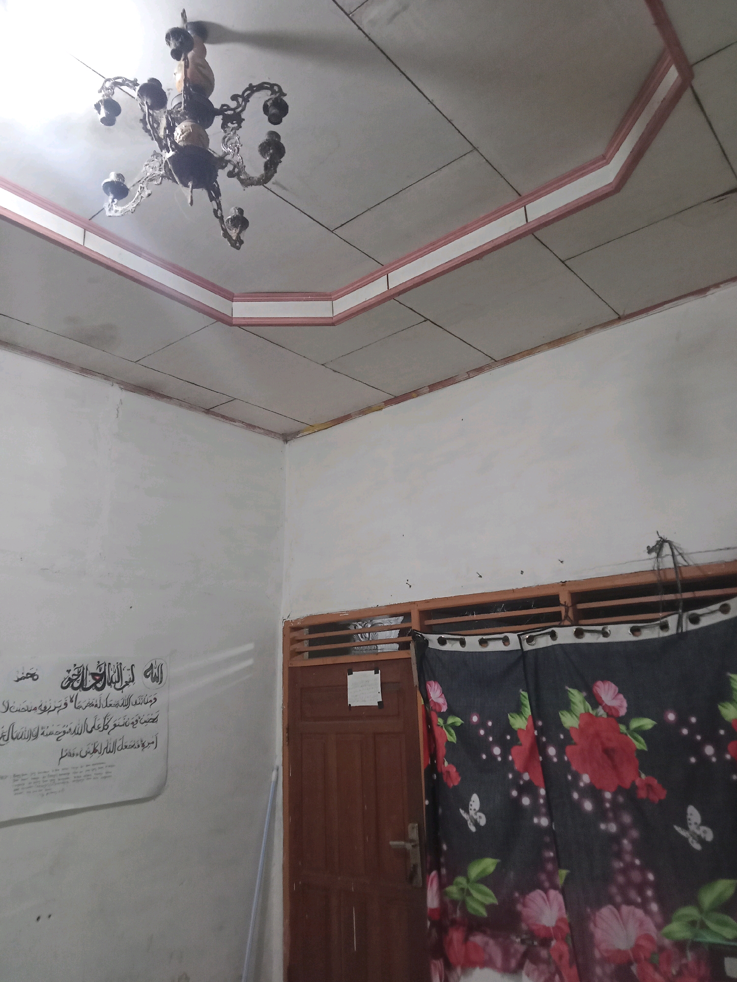 The image shows an interior view of a room, likely in Sukomananti, Indonesia. A vintage-style chandelier hangs from the center of the white ceiling, which has a decorative pink and white trim. The walls are painted white. On the left wall, there is a framed piece of calligraphy in Arabic script. To the right, there is a wooden door with a silver handle and a window with horizontal wooden slats above it. A dark curtain with a floral pattern of red and pink roses and white butterflies hangs in front of the window. The floor is not visible. The room appears to be a residential interior, possibly a living room or bedroom.