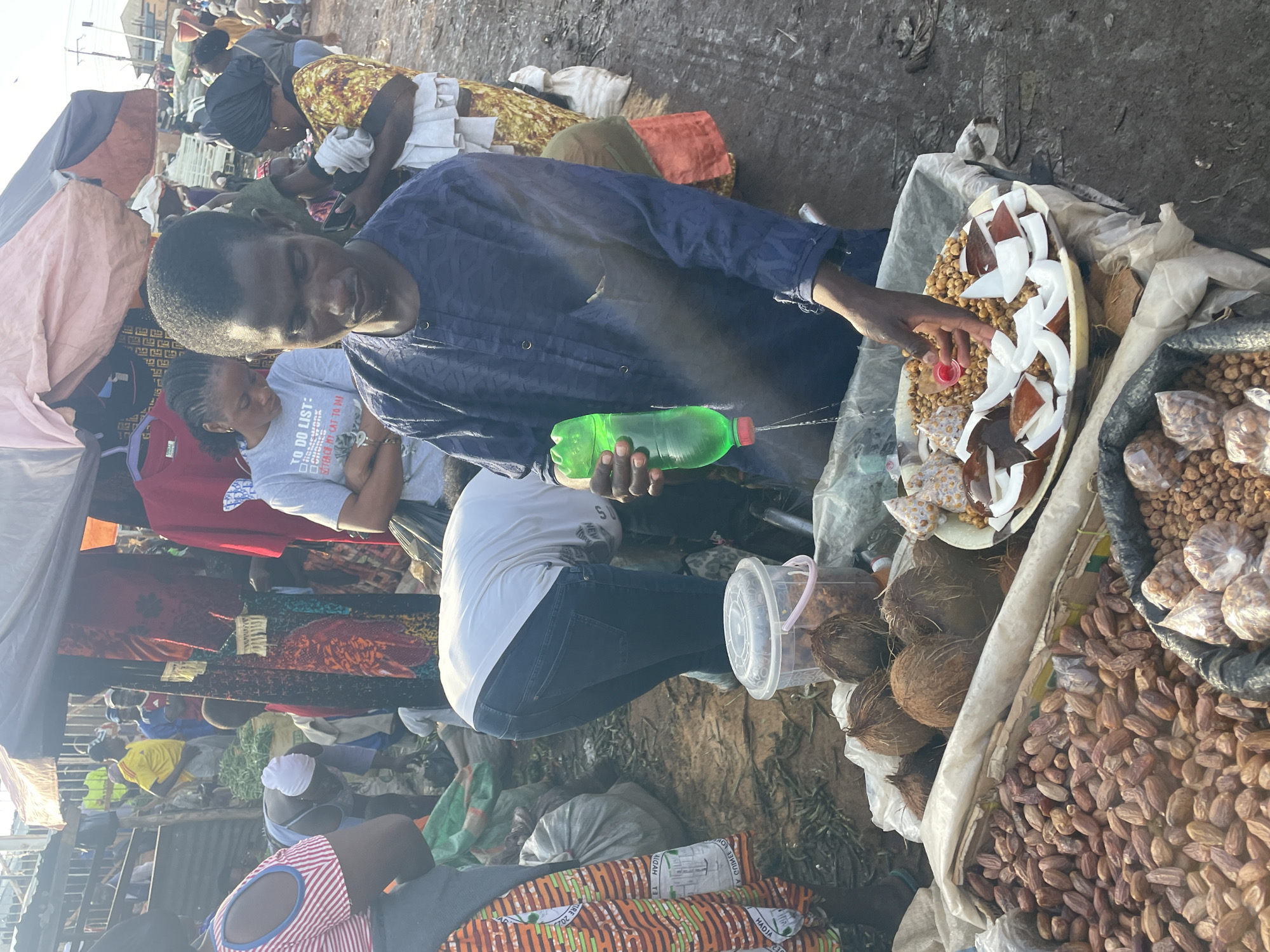 This image captures a bustling market scene in Gura Top, Nigeria. In the foreground, a man wearing a dark blue patterned tunic is pouring a green liquid from a plastic bottle into a small red cup. He is standing behind a table laden with various goods. To his left, a large pile of dried dates is visible, with coconuts and smaller bags of nuts also on display. To his right, a plate contains chopped coconut pieces and what appears to be small fried dough balls or nuts.
Behind the man, several other people are visible. A woman in a yellow and black patterned dress is holding a baby. Another woman in a light-colored shirt with a "To Do List" graphic is looking down. The market appears to be outdoors, with stalls covered by tarps. The ground is dirt and debris. The overall impression is one of a vibrant, active marketplace with a focus on food vendors.
