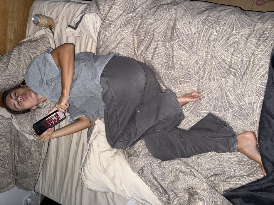 The image portrays an indoor bedroom scene in Warsaw, Poland. A single individual, appearing female, is depicted lying on her left side on a bed. She is attired in a light grey short-sleeved t-shirt and dark grey sweatpants, with her bare feet positioned towards the bottom of the frame. Her head is resting on a pillow featuring a grey-brown abstract leaf or feather pattern. Her eyes are partially closed, and her lips are puckered in a distinct pout. She is holding a black smartphone horizontally with both hands, orienting it towards her face. The back of the smartphone case displays the text "MÁS AMOR POR FAVOR" in red and white lettering. To the right of her head, an upright, golden-colored aluminum beverage can, appearing to be Coca-Cola Zero, is placed on the bed's light beige fitted sheet. The bed's bedding includes two patterned pillows, a patterned duvet, and a light beige fitted sheet, with additional lighter and darker blankets visible. A wooden headboard is partially visible to the right of the individual's head, and a light-colored wall with a white power outlet and a plugged-in cord is visible behind the bed on the left.
