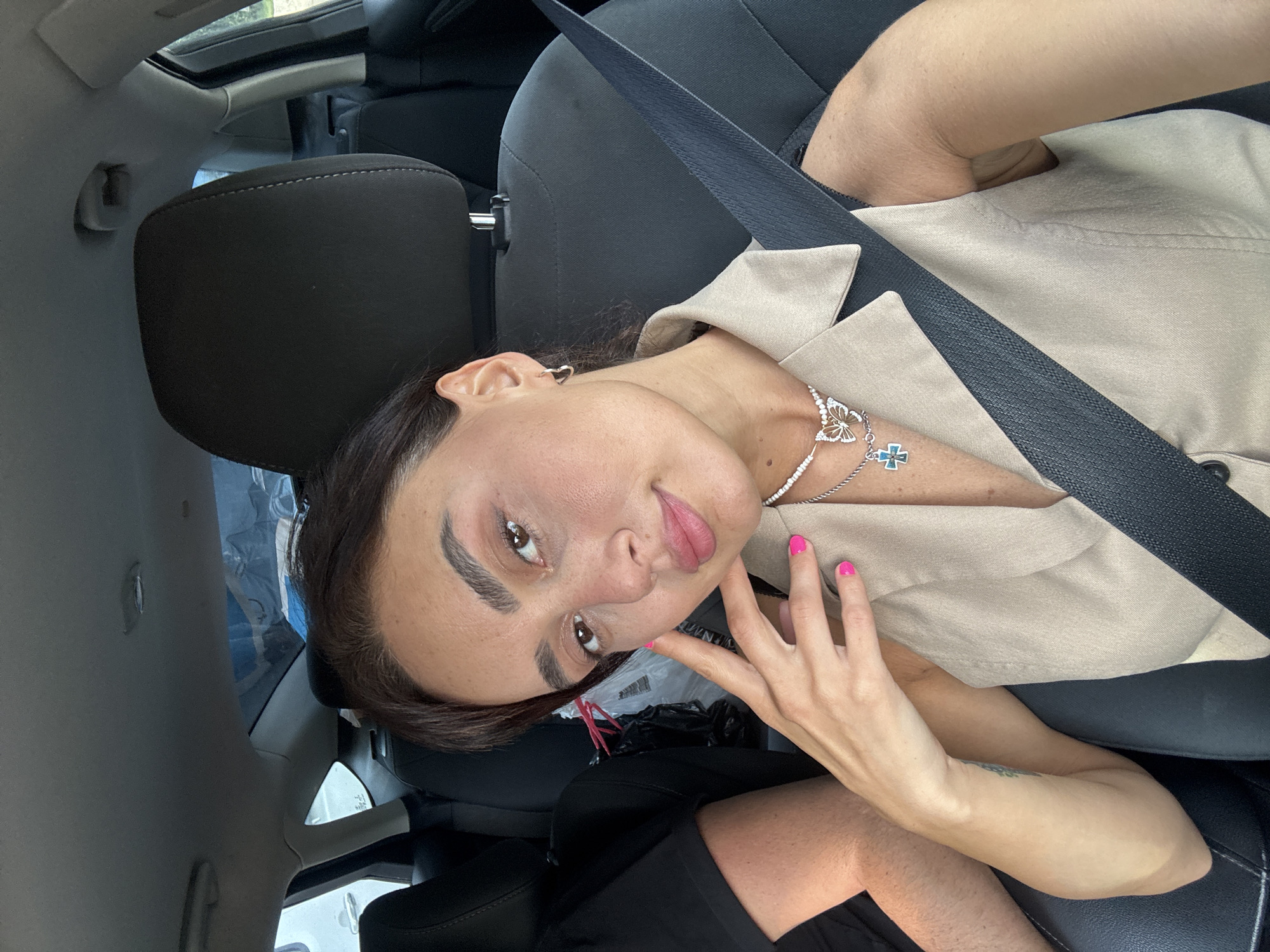 A woman is sitting in the passenger seat of a car, looking at the camera. She has dark hair and is wearing a beige sleeveless shirt and a necklace. Her fingernails are painted bright pink. A seatbelt is visible across her chest. The interior of the car is dimly lit, with the back seat and headrests visible behind her. The image appears to be a selfie taken inside a vehicle. The location is not specified to be Tbilisi, Georgia.