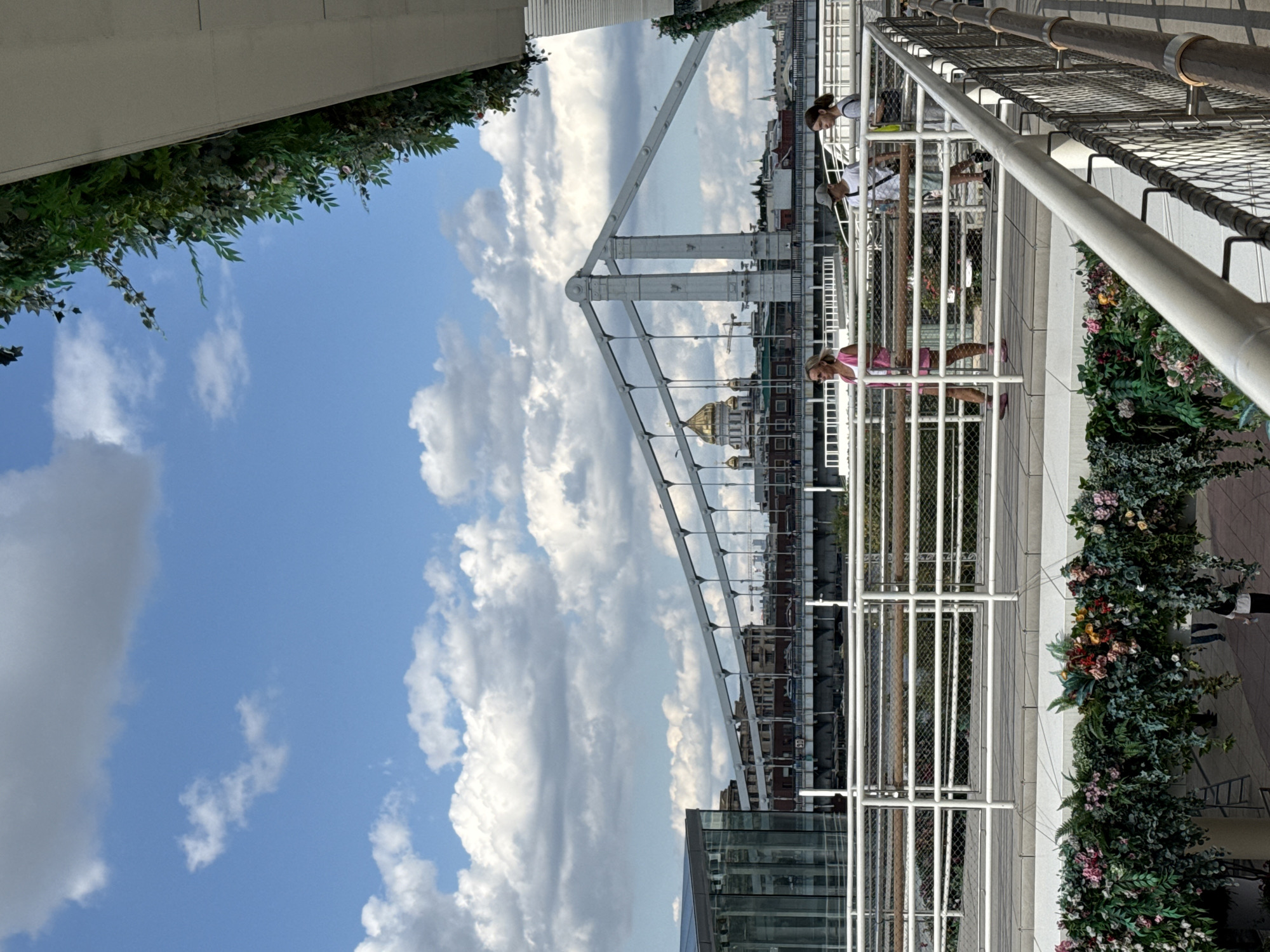 The image captures a view from an elevated walkway in Moscow, Russia, overlooking the Krymsky Bridge and the golden dome of Christ the Savior Cathedral in the background. The walkway is adorned with lush greenery and floral arrangements. In the foreground, three individuals are walking along the walkway. One person, wearing a pink outfit, walks ahead, while two others, one in a grey cap and white t-shirt and another in a light grey t-shirt, follow behind. The sky is a clear blue with scattered white clouds. To the left, a modern glass building is visible. The Krymsky Bridge, a suspension bridge, spans the Moscow River, with its distinctive white towers and cables. The overall scene depicts a pleasant day in an urban setting, with a blend of modern architecture, historic landmarks, and decorative natural elements.
