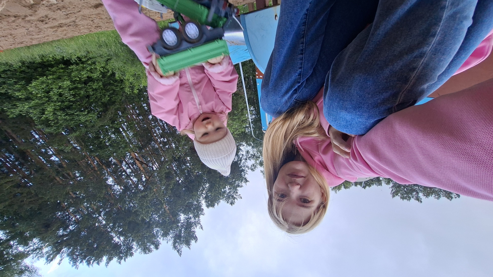 An adult female and a young child are observed outdoors from a low-angle perspective, consistent with a camera held by the adult. The adult, positioned on the left, has blonde hair, light skin, and wears a pink hooded sweatshirt and blue jeans. She is looking directly into the camera. A light blue and brown structure, possibly part of a playground slide, is partially visible behind her to her right. The child, situated on the right, wears a light pink full-body suit and a light-colored knitted beanie. The child is holding and manipulating a green and dark-colored toy truck with both hands, focusing on the toy. The ground surface where the child is playing appears to be sand or dirt. The background consists of numerous tall pine trees and other green foliage under an overcast or cloudy sky. There are no discernible landmarks or textual elements in the image to confirm the specific location as Chaikovsky, Russia.
