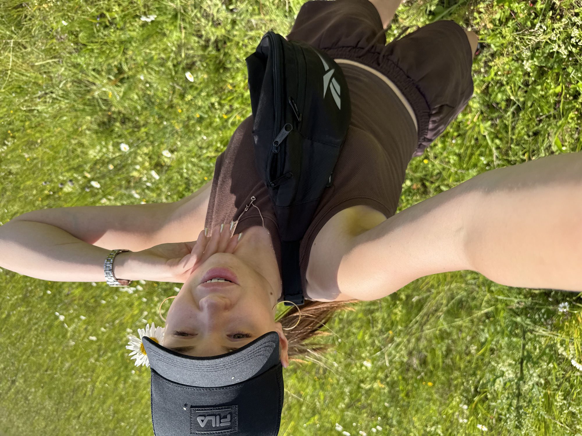 A young woman with light skin and brown hair lies on a grassy field speckled with small white flowers, facing upwards and towards the camera. She is wearing a brown crop top, brown shorts with white trim, a black fanny pack, and a black baseball cap adorned with a white daisy. A silver watch is visible on her left wrist, and she wears large gold hoop earrings. Her expression is relaxed and she lightly touches her face with her right hand. The scene appears to be outdoors on a sunny day, with dappled sunlight creating shadows on the grass. The overall mood is peaceful and casual. Visible text includes "Reebok" on the fanny pack and "FILA" on the baseball cap. The provided location context is "Гаревая, Russia."